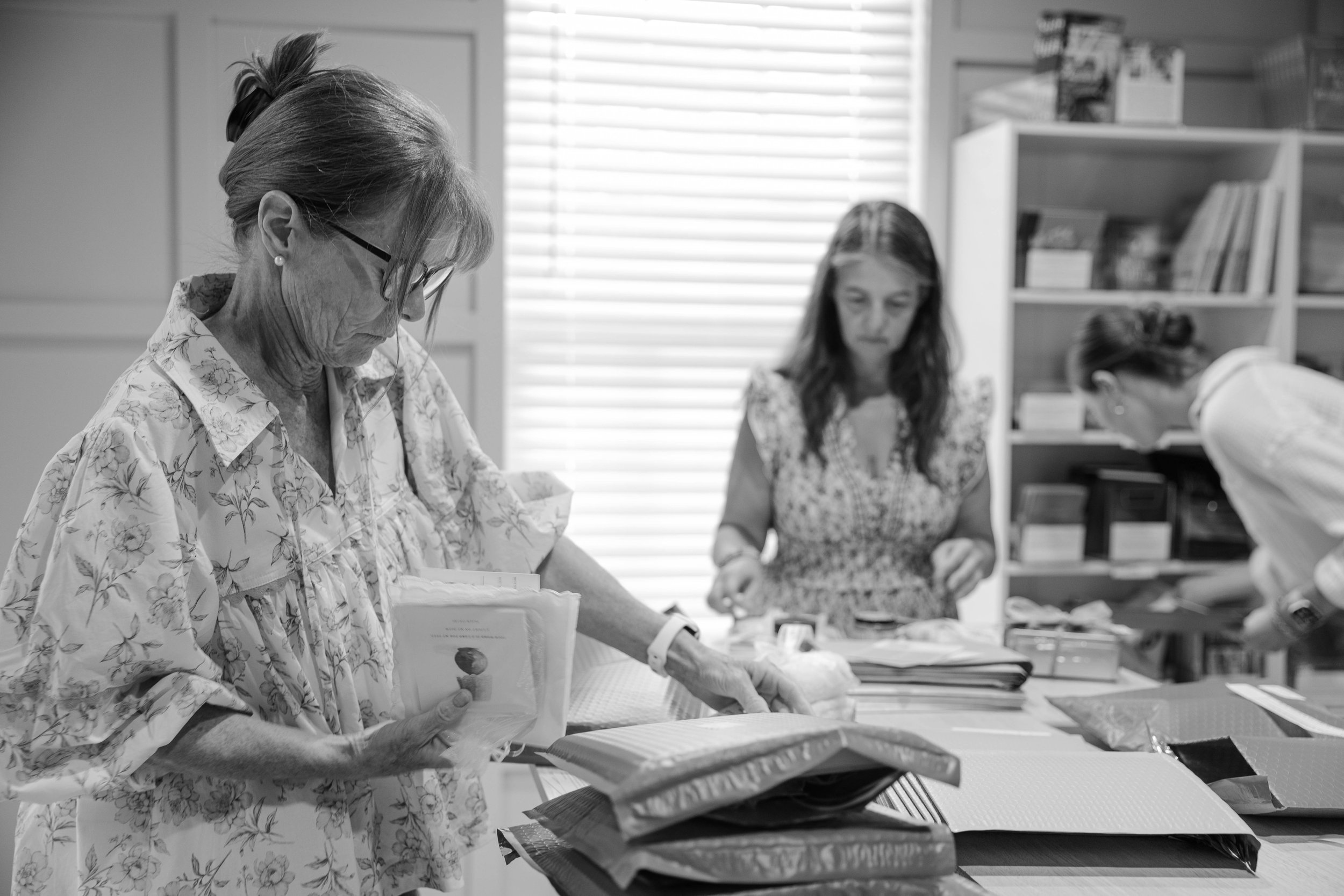 An elderly woman with glasses and earrings, wearing a floral blouse, is working at a table with books and documents. Behind her, two younger women are also engaged in work, with shelves filled with books and supplies in the background.