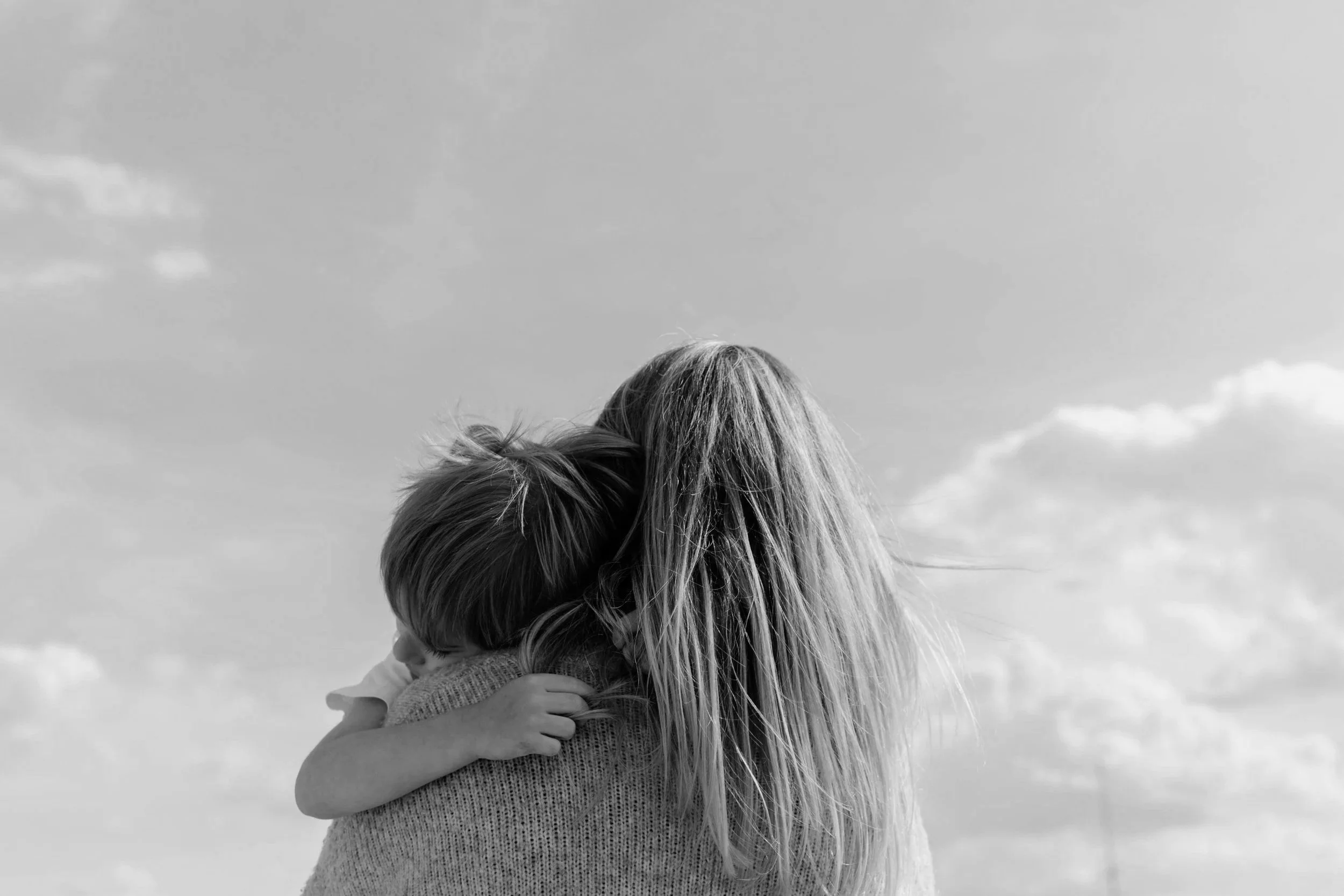 A person with long hair hugging a child, both facing away from the camera, outdoors with a cloudy sky.