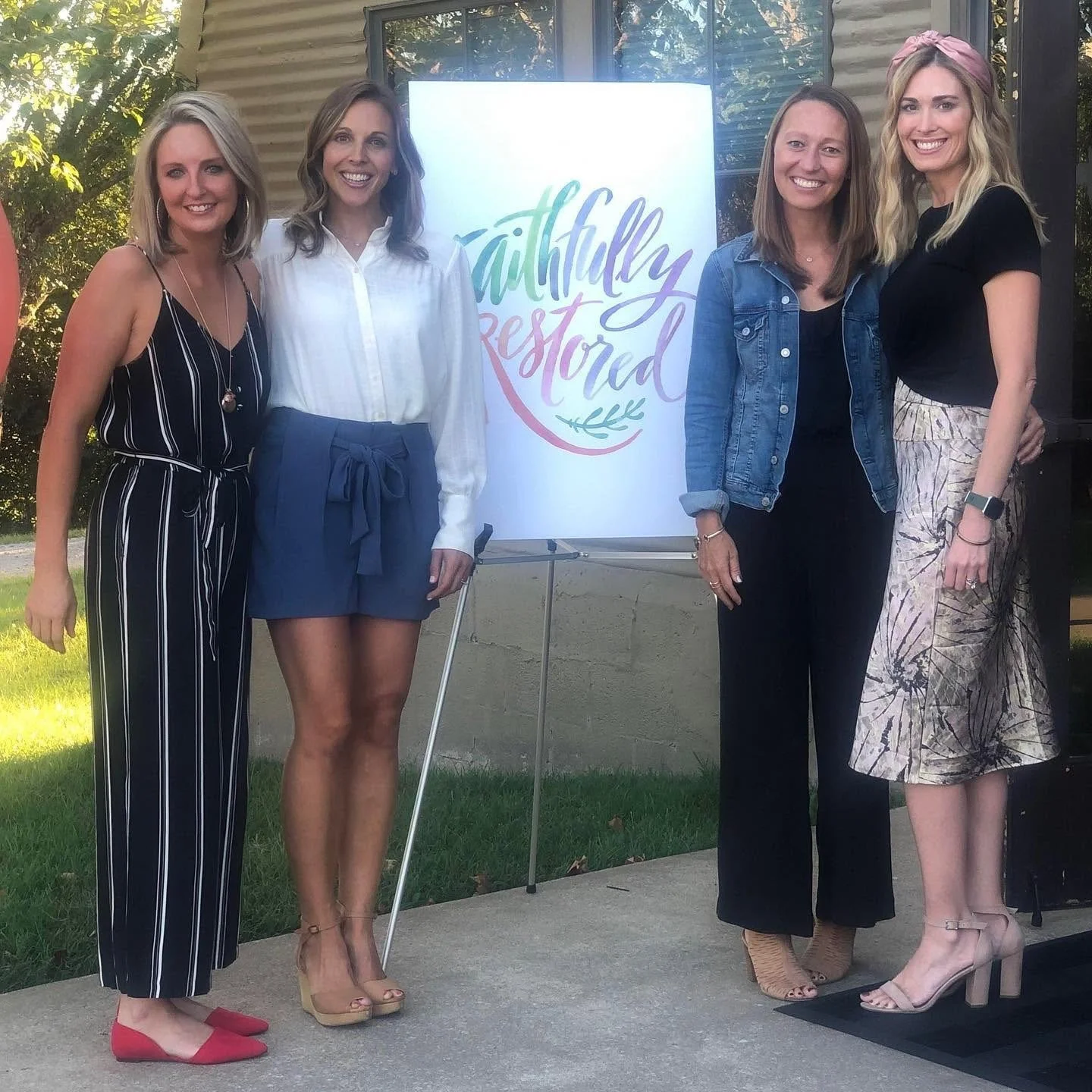 Four founding women are standing outside in front of a sign that reads 'Faithfully Restored.' They are smiling and dressed in summer outfits.