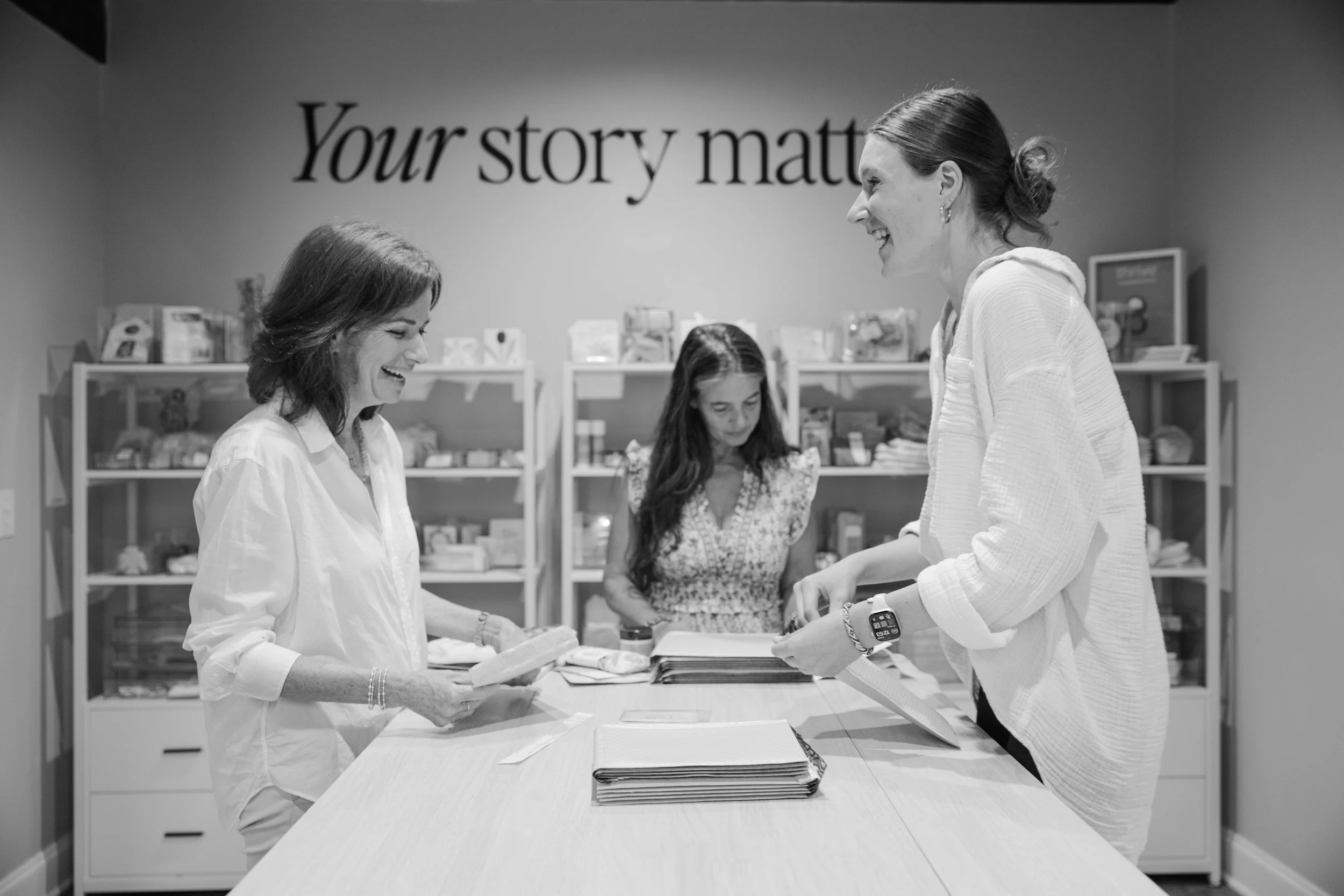 Three women in a meeting room looking at documents on a table, smiling and engaging in conversation. The background features shelves and a sign that reads 'Your story matter'.