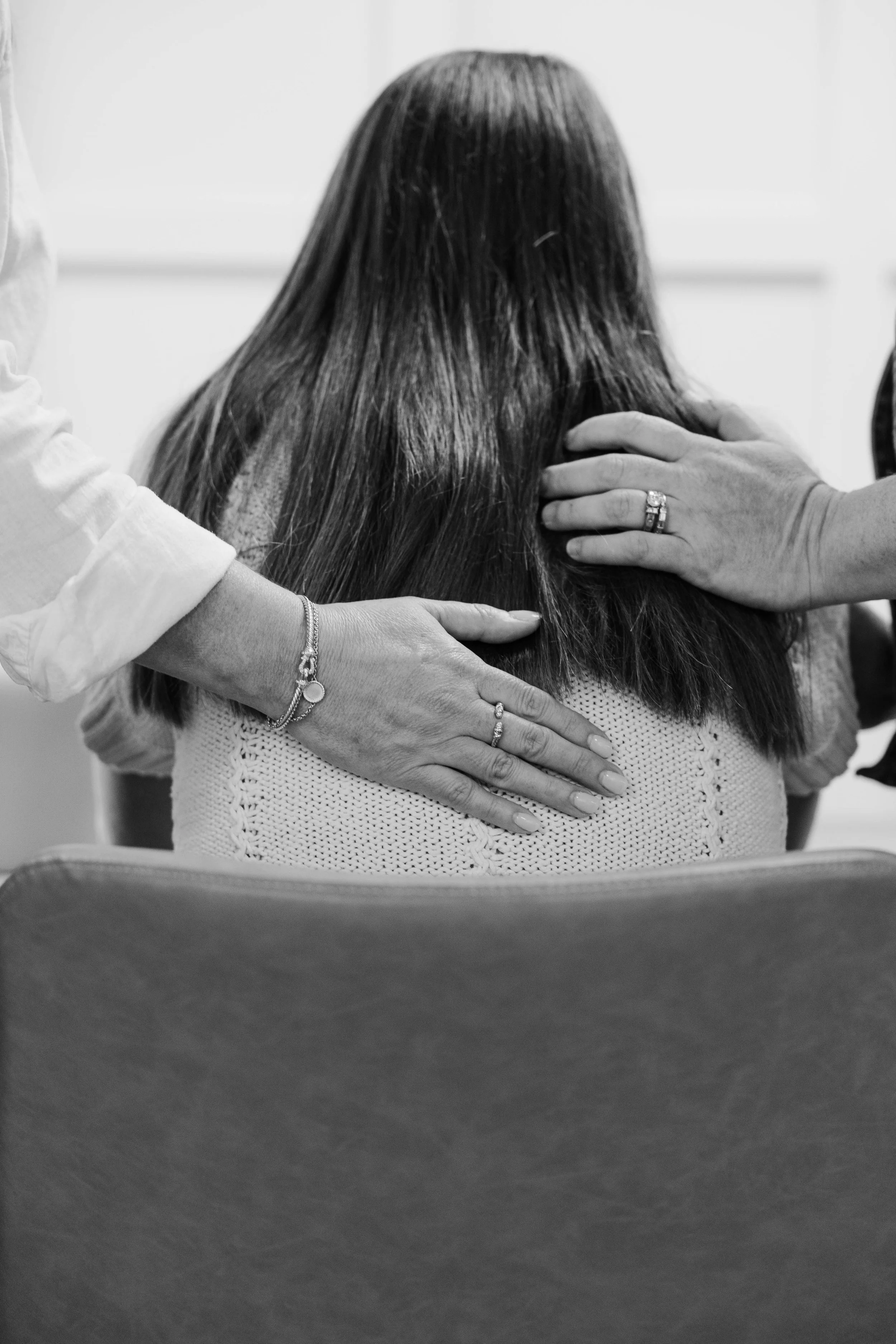A person with long hair is sitting with their back to the camera. Two other people are touching the person's shoulders and upper back, one on each side. The person on the right is wearing rings and a bracelet.