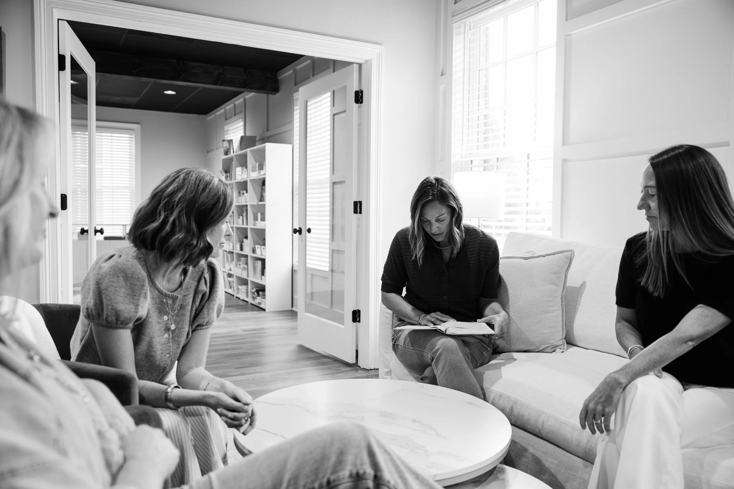 A group of women sitting and engaging in a discussion around a table in a bright, cozy living room with large windows, white walls, and bookshelves.