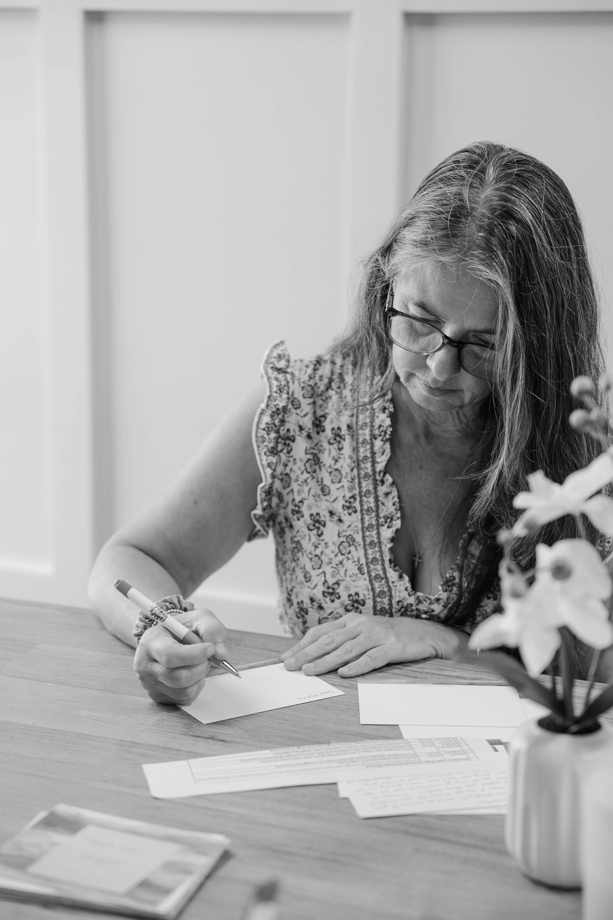 A woman with glasses and long hair, writing on a piece of paper at a wooden desk, with papers and a potted plant with flowers in the foreground.