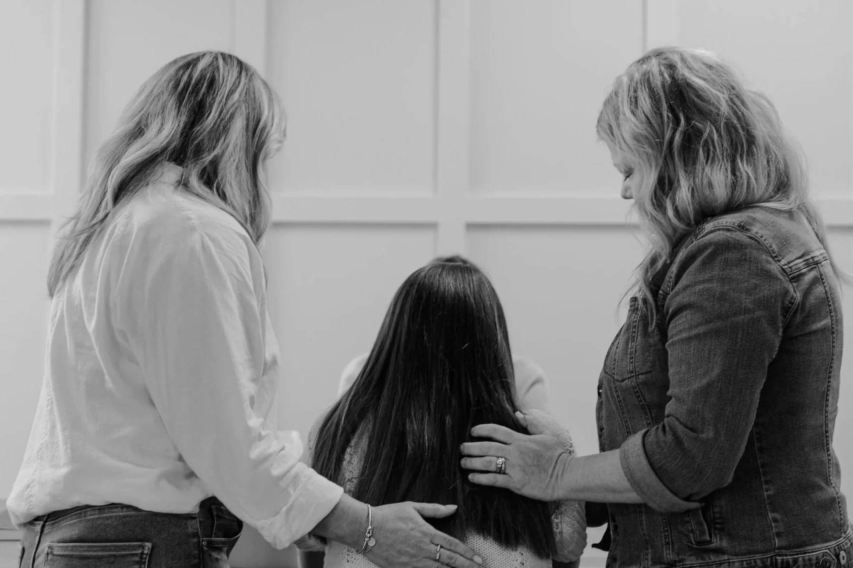 Two women and a girl with long dark hair facing each other in prayer or support, with the women’s hands gently placed on the girl’s shoulders and back, in a grayscale photo.
