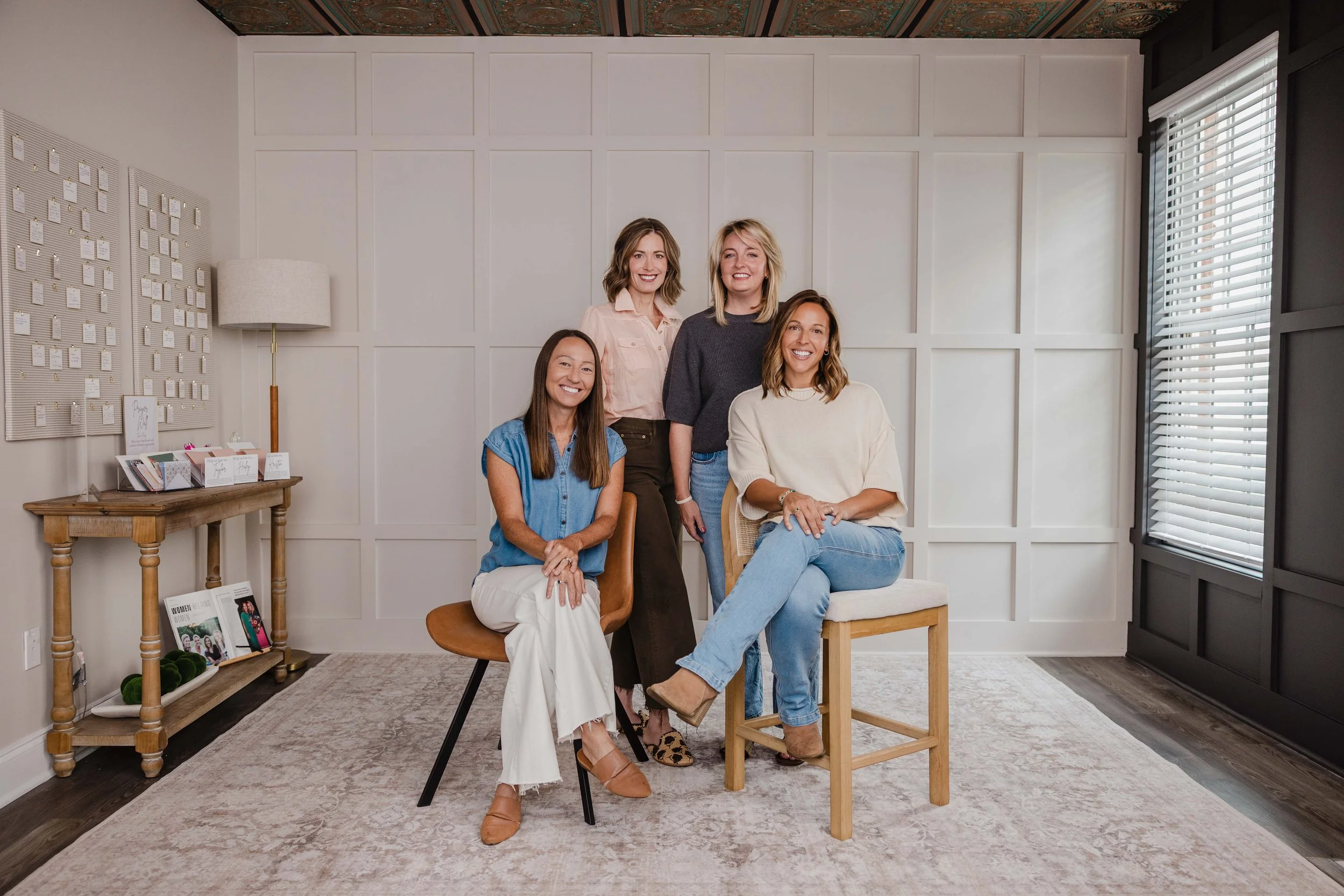 Five women in a cozy, well-decorated room, posing happily for a photo. The background features a white-paneled wall, a window with blinds, a lamp, and a small wooden table with magazines and decorative items.