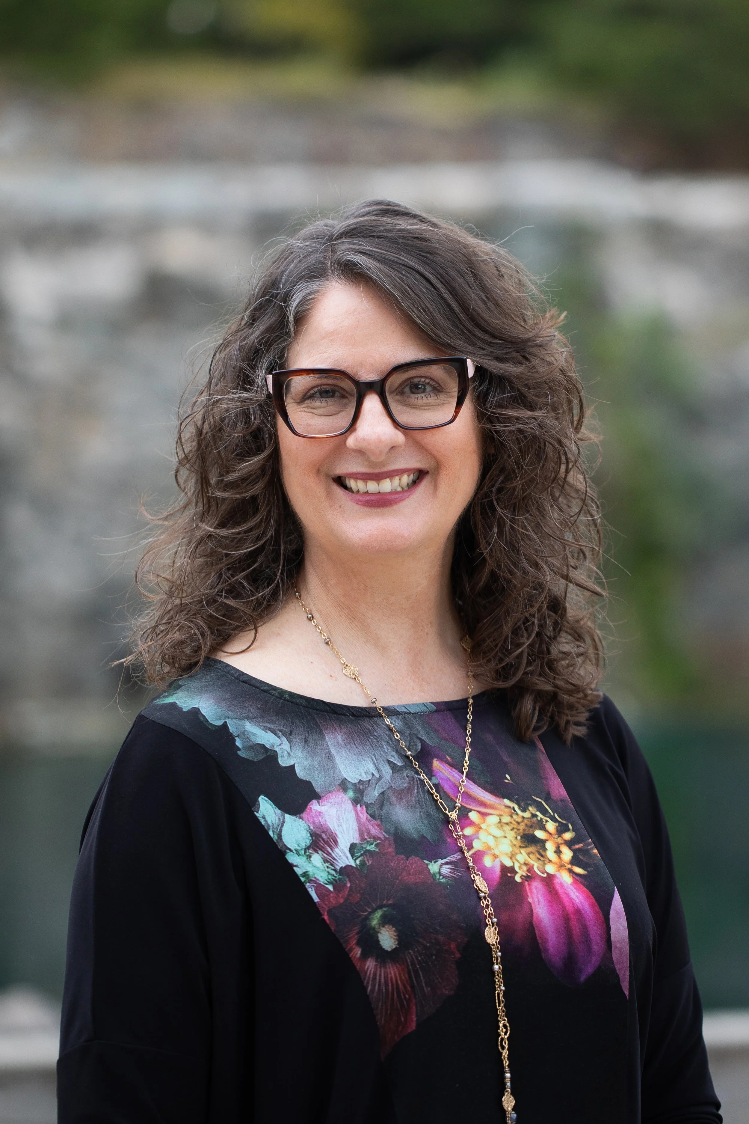 Black and white photo of a woman with long, curly hair wearing glasses and a patterned shirt, smiling and sitting outdoors.