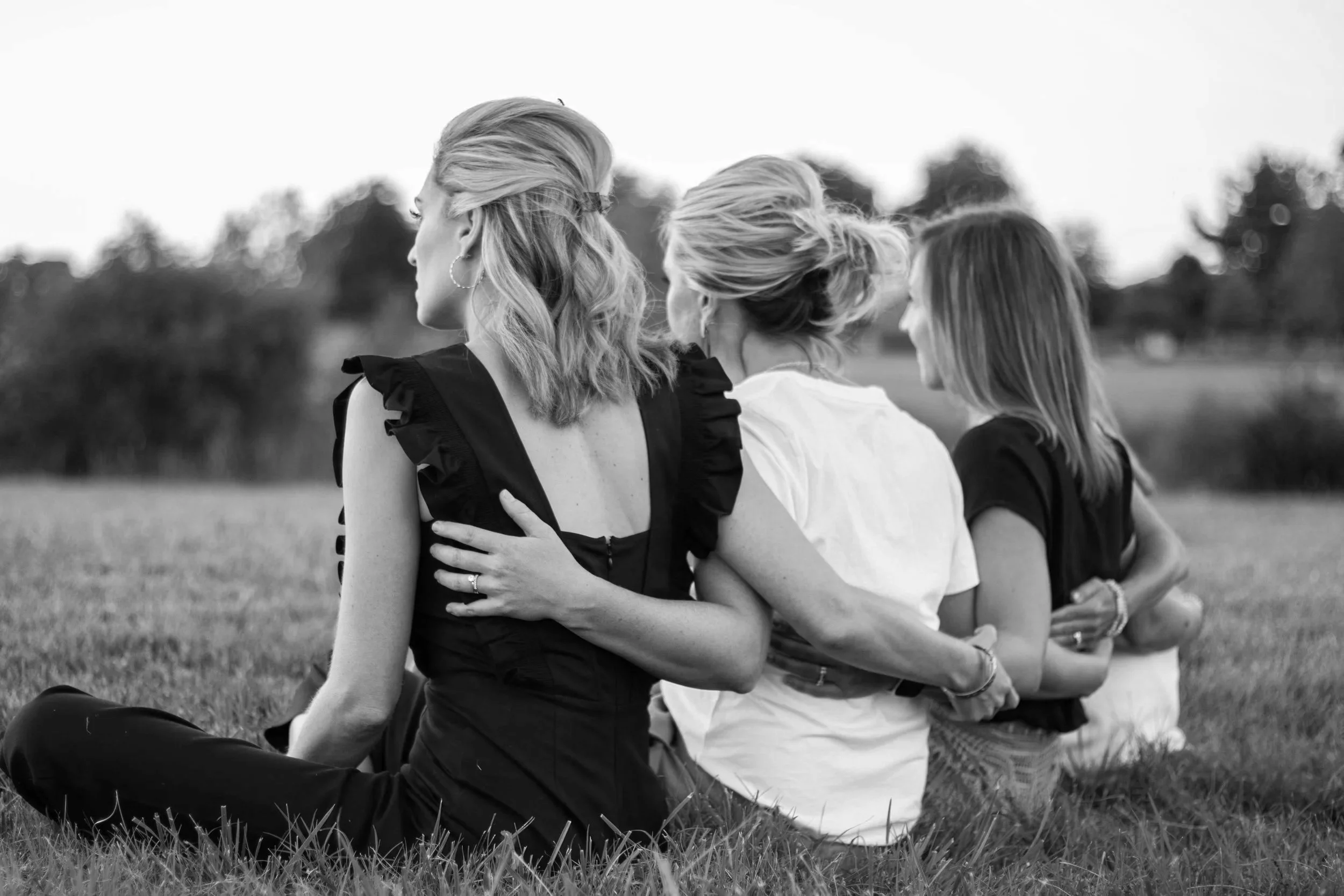 Four women sitting on grass outdoors, embracing and appearing to enjoy each other's company.