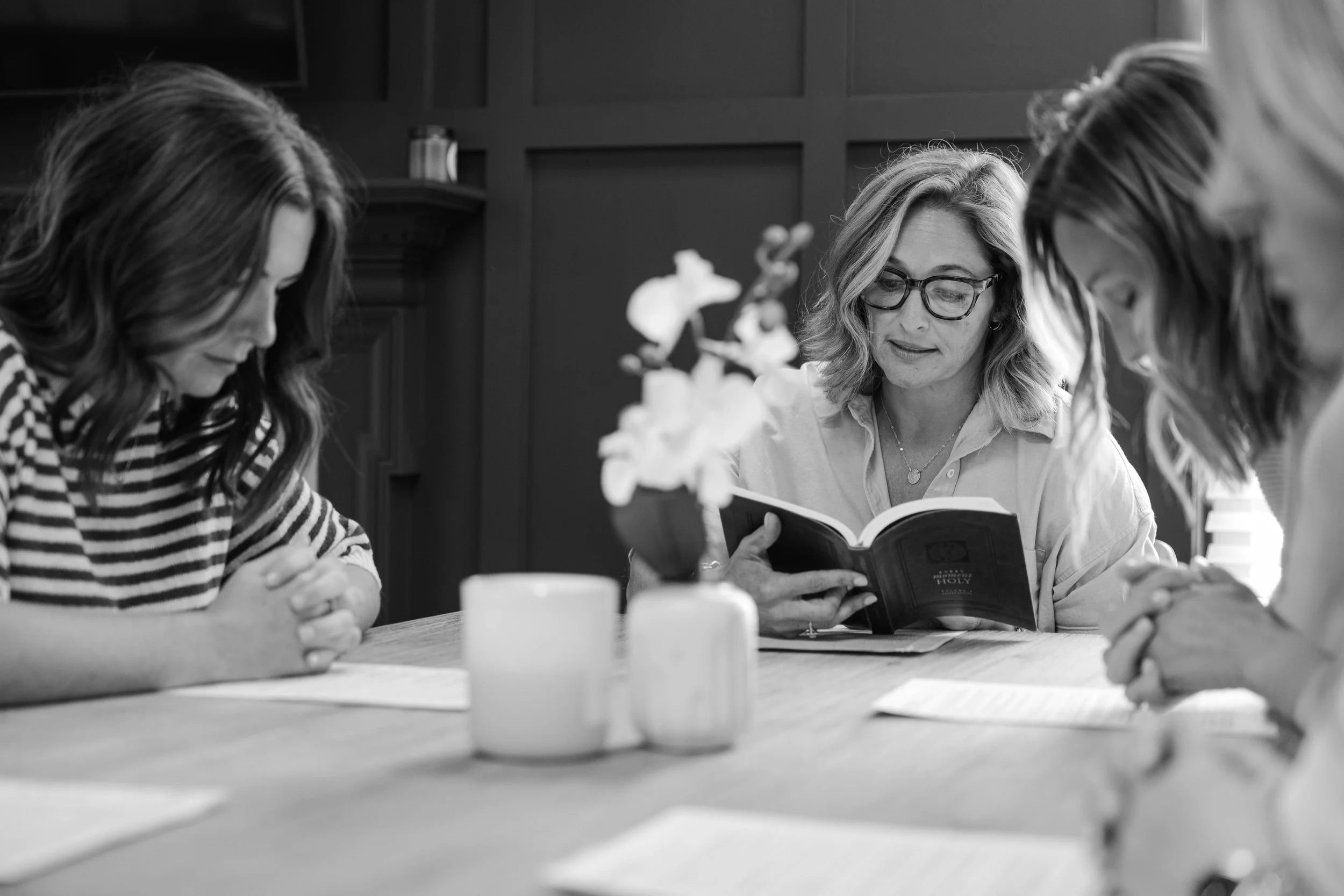 Three women sitting at a table, engaged in reading a book together, with a flower vase in front of them, in a room with dark paneled walls.