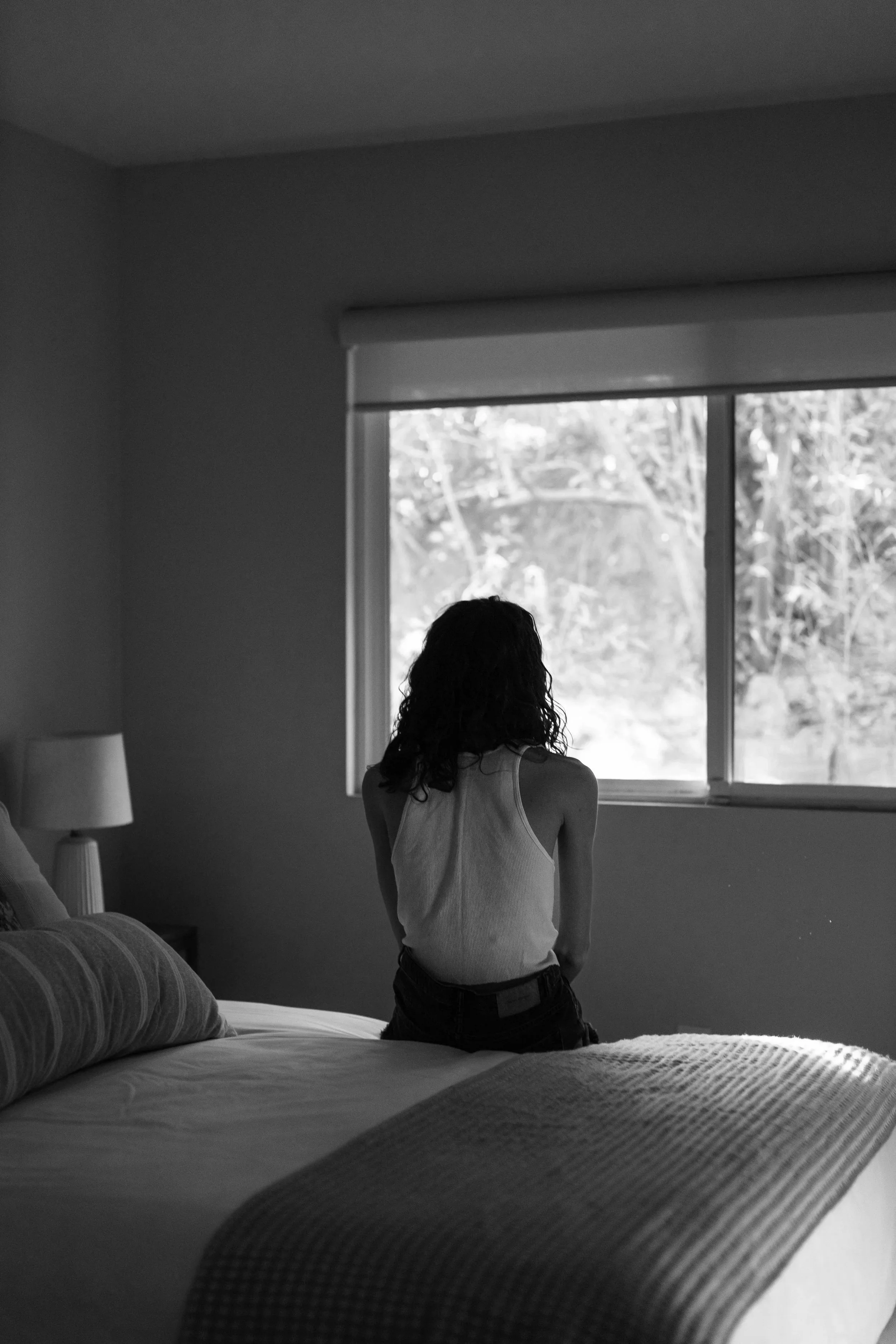 A woman with curly hair sitting on a bed, facing a window that looks out to trees and natural scenery. The room has muted lighting and a side table with a lamp.