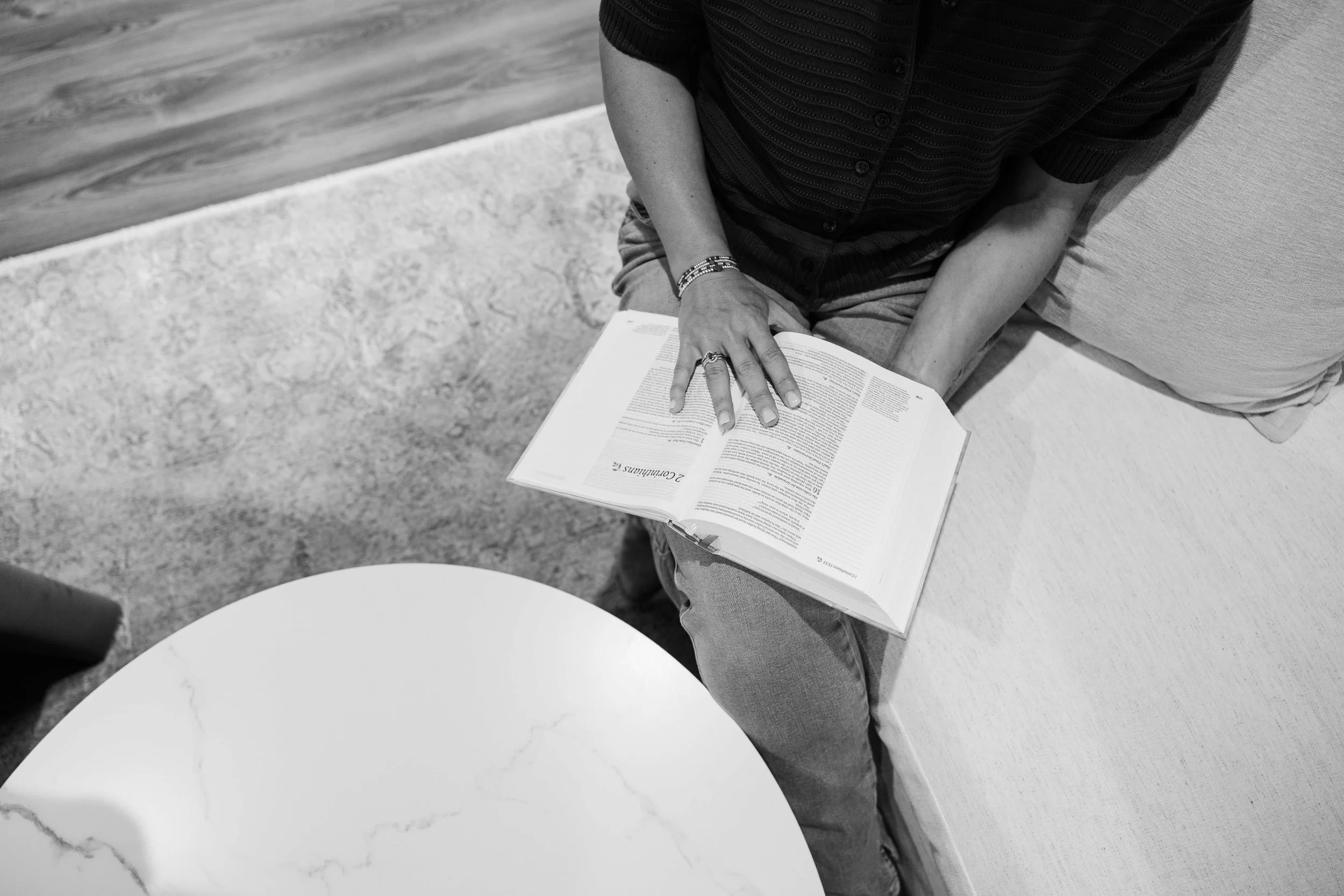Person sitting on a sofa reading an open book, with part of their arm and hand visible, wearing a dark striped shirt and jewelry.