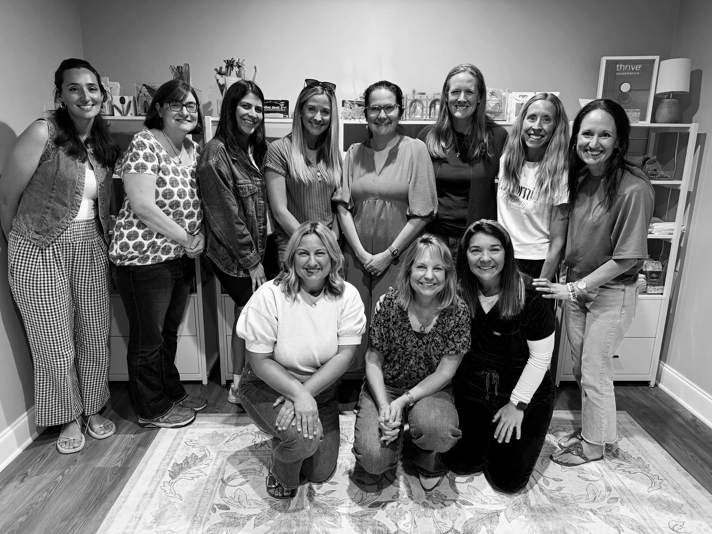 Black-and-white photo of a group of women in a room, some standing and some kneeling, smiling at the camera, with shelves and decor in the background.