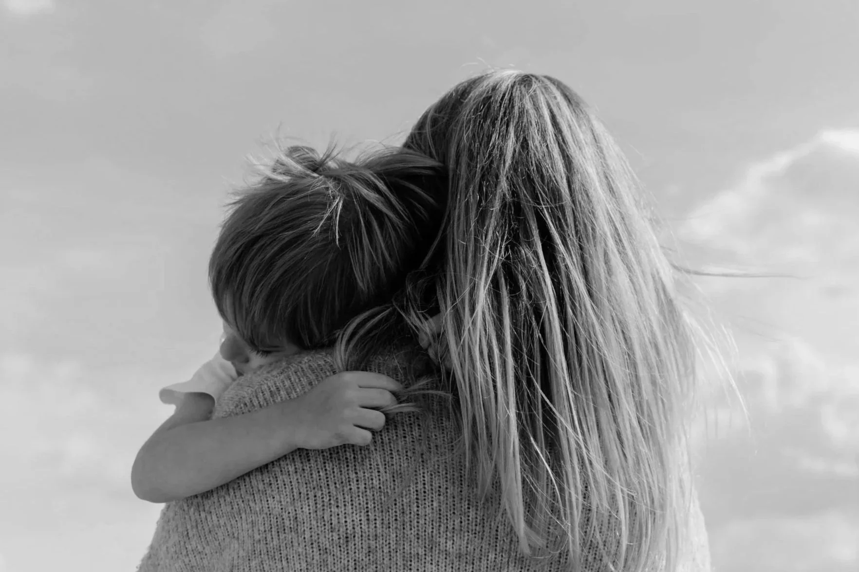 Black and white photo of a woman hugging a young child outdoors against a cloudy sky.
