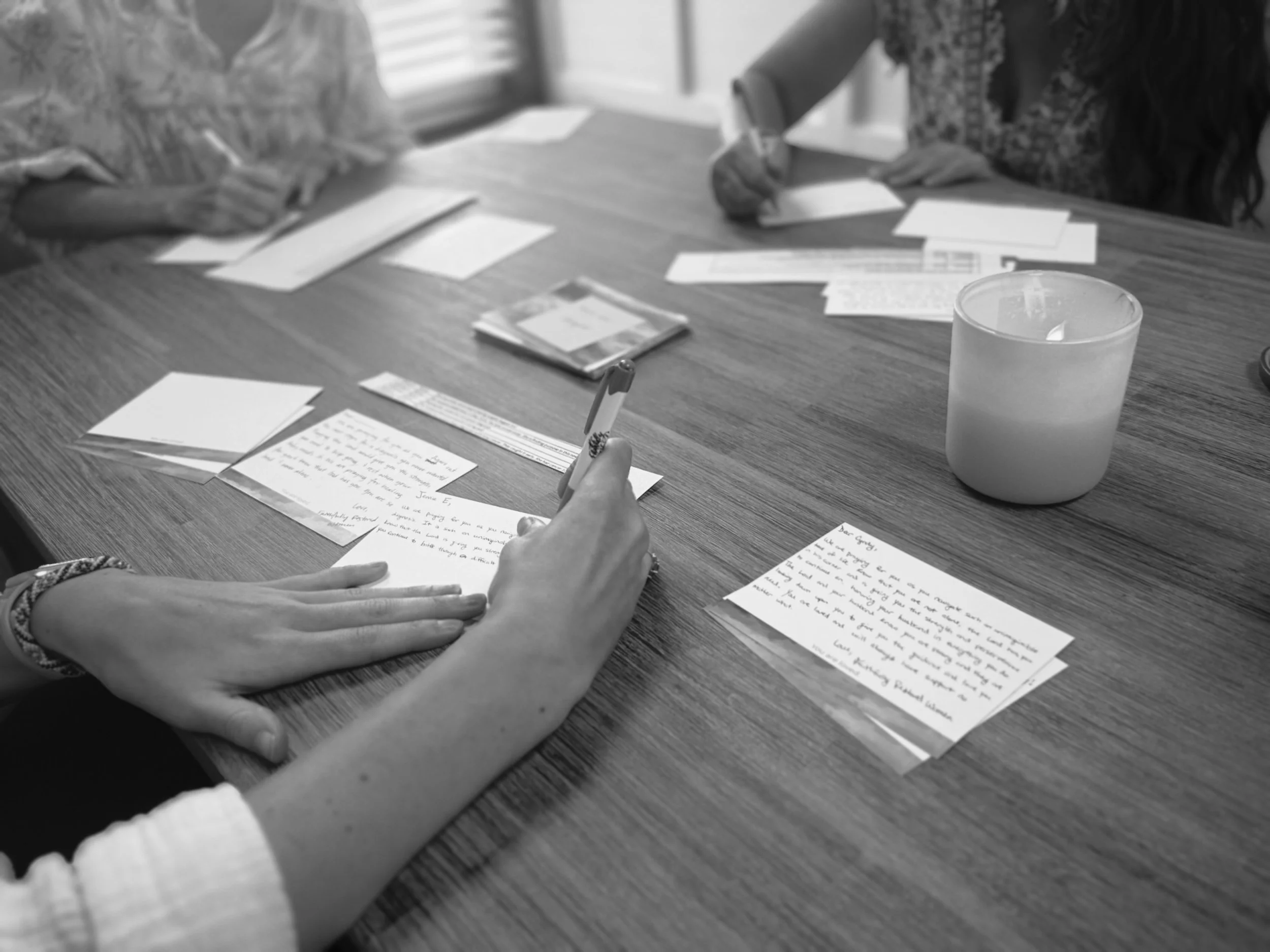 Two people sitting at a table writing letters or cards, with a candle, papers, and envelopes on the table.