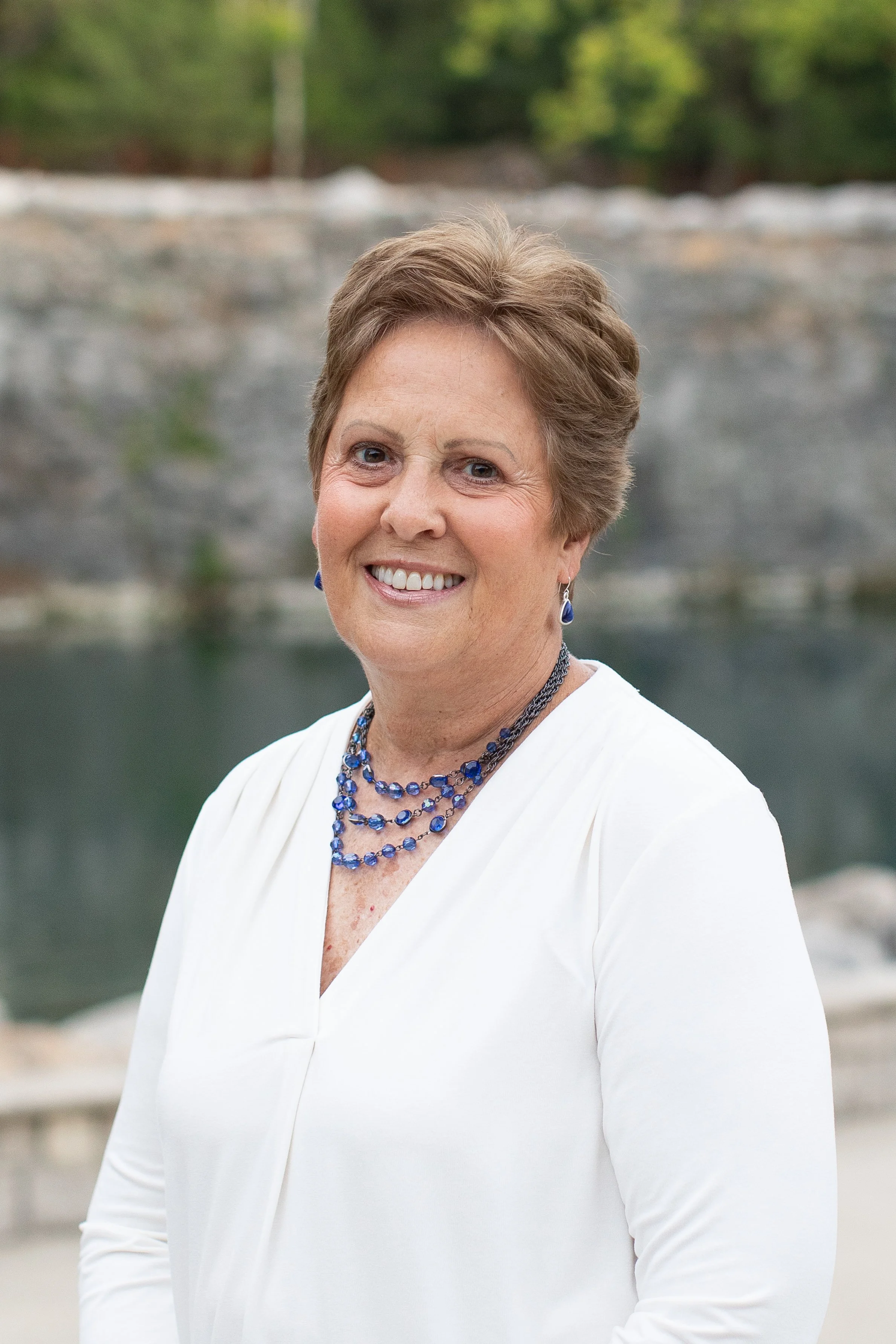 A smiling woman wearing a dark dress, pearl necklace, and earrings, standing outdoors in front of blooming hydrangeas.