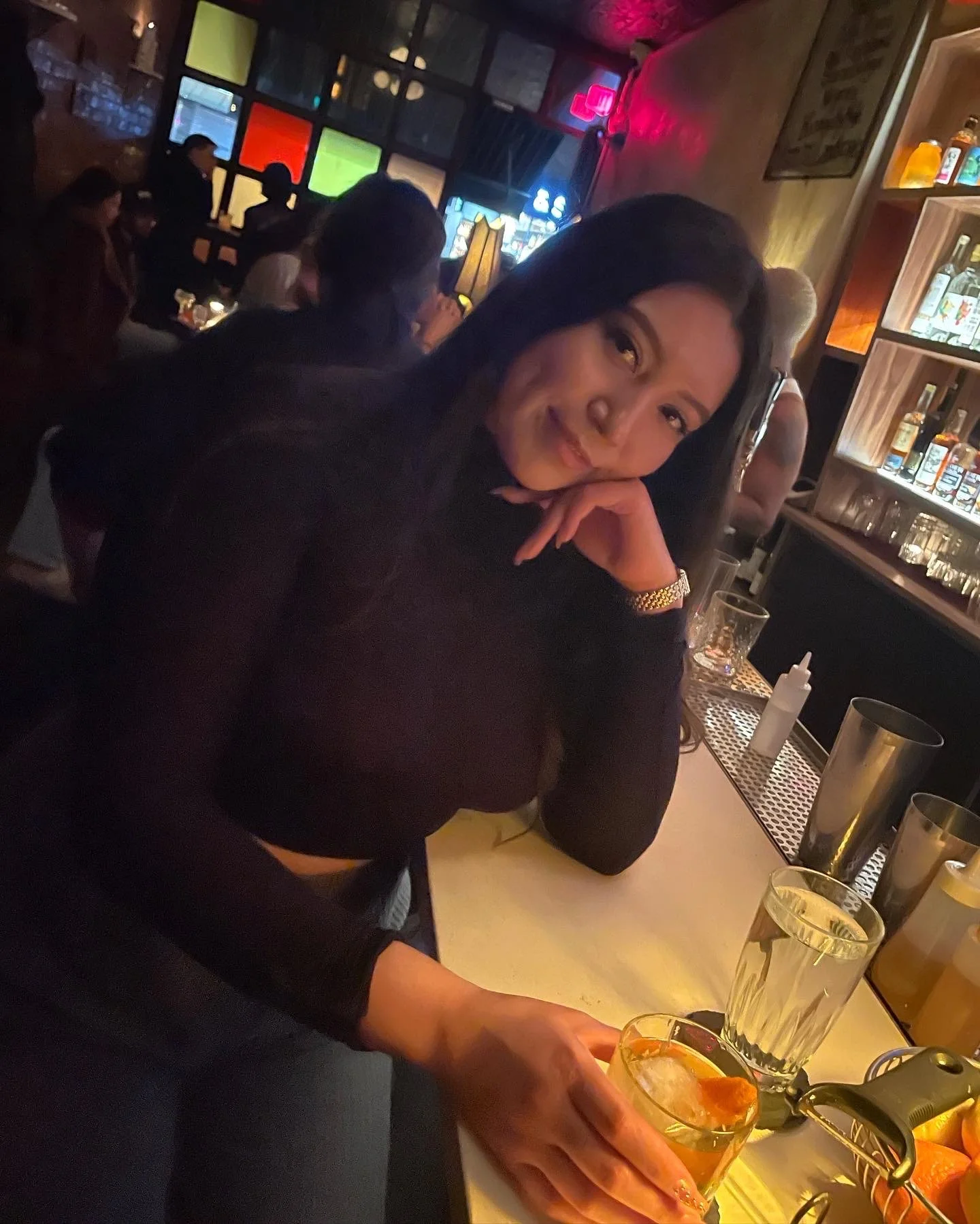 A woman in a black shirt sitting at a bar counter, holding a drink with ice and orange slices. The background shows other people seated, a bar shelf with bottles, and colorful display panels on the wall.