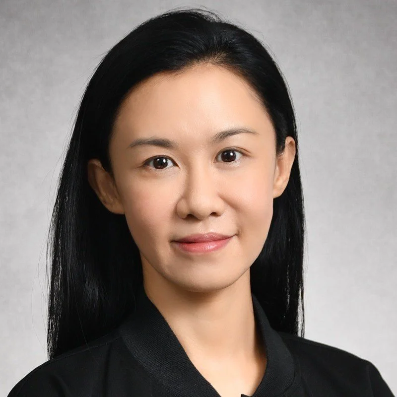 A woman with straight black hair and a slight smile, wearing a black top, against a plain gray background.