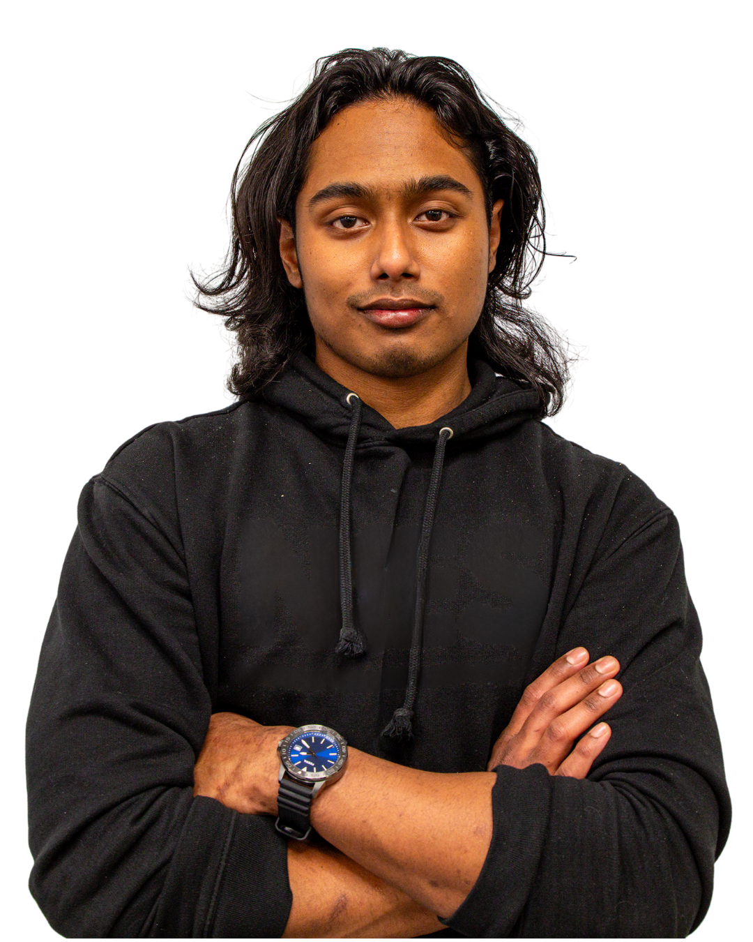 A young man with medium skin, long black hair, and wearing a black hoodie crossing his arms, looking directly at the camera, with a black background.