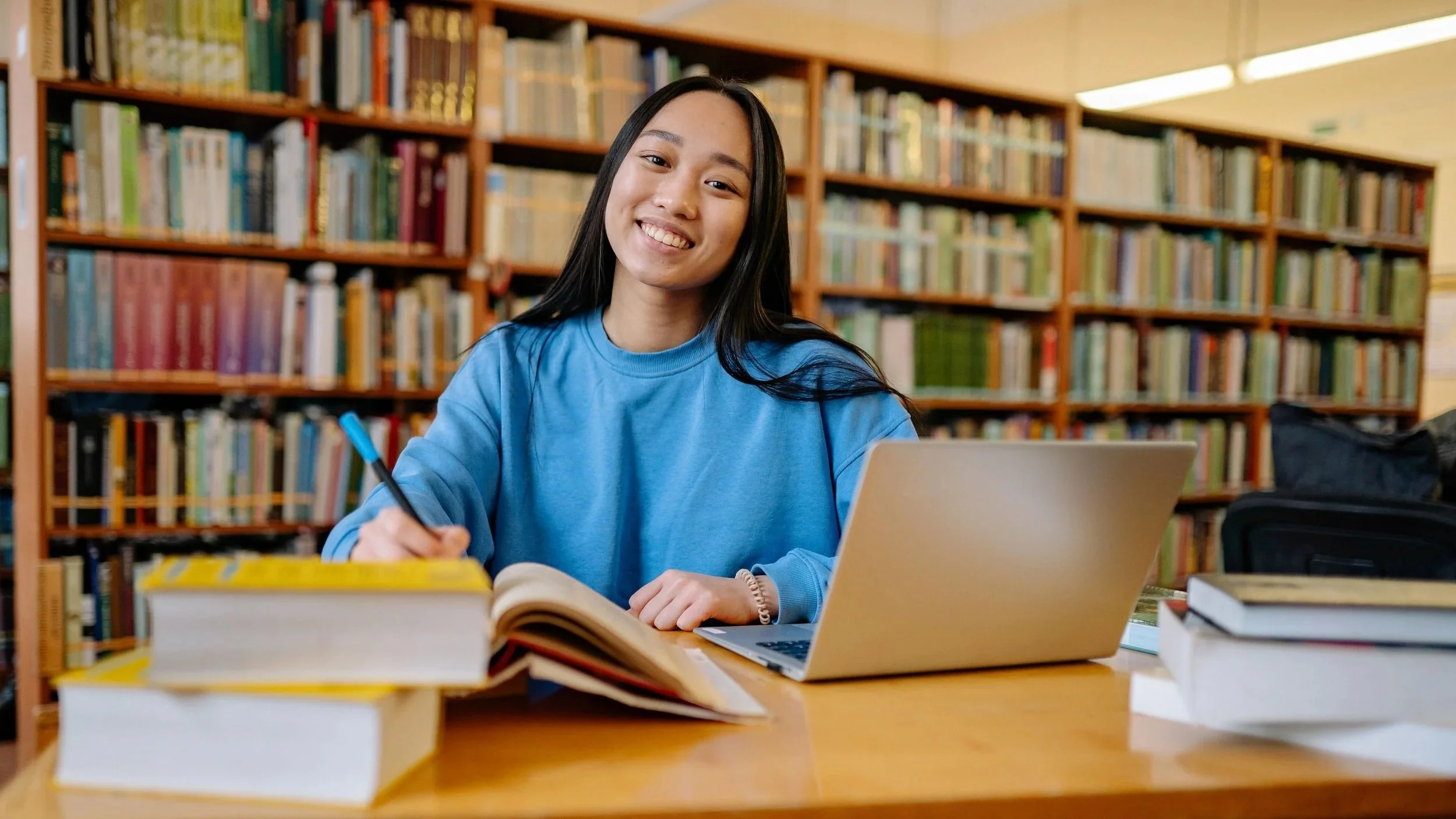 A young woman with long black hair, wearing a blue sweatshirt, sitting at a library table with books, a laptop, and an open notebook. She is smiling and appears to be studying or working.