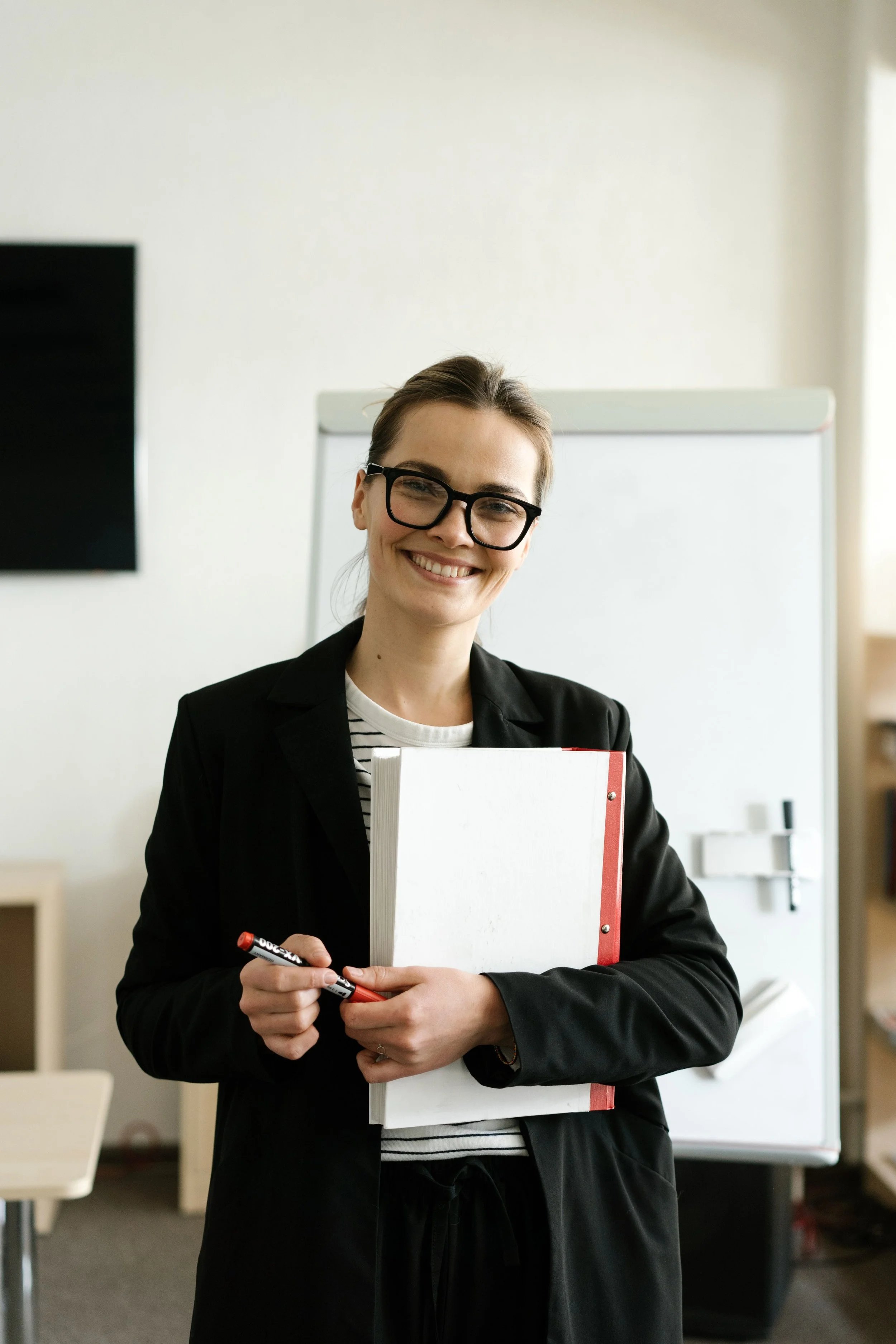 Smiling woman with glasses holding a whiteboard and a marker in a classroom or office setting.