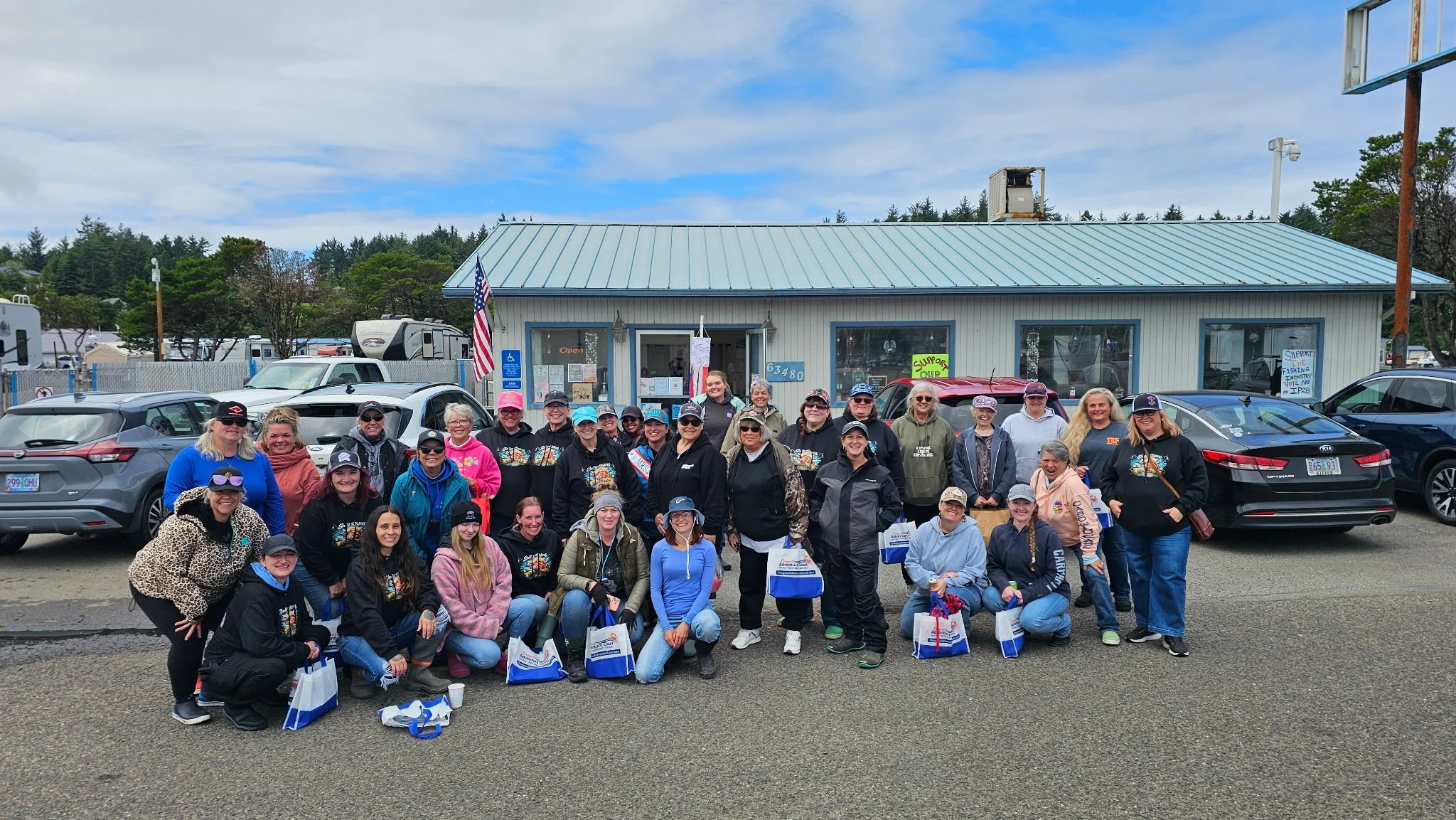 A large group of people posing together in a parking lot in front of a light blue building with signs, flags, and cars parked nearby.