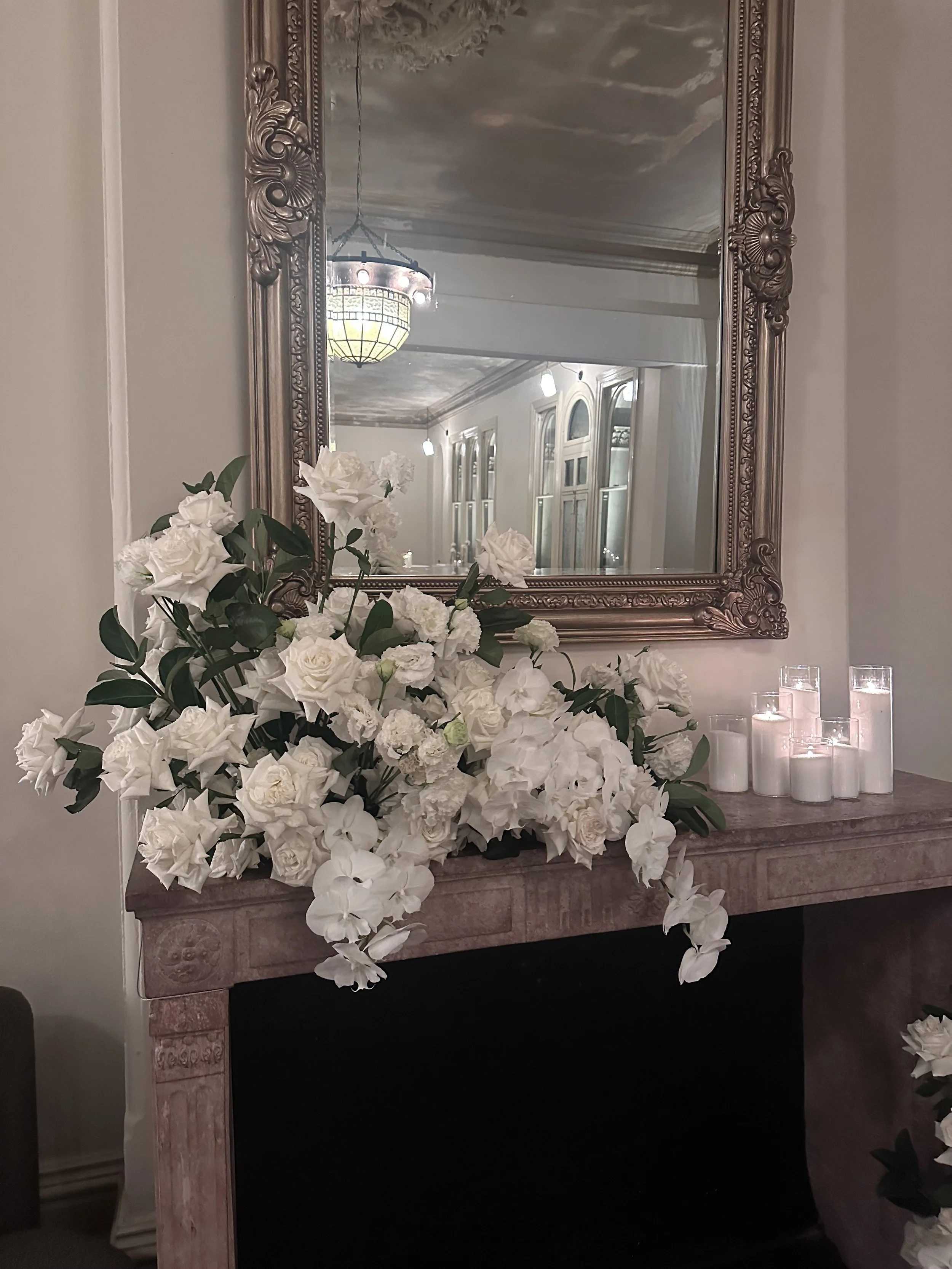 A large floral arrangement of white flowers on a marble fireplace mantel with a decorative mirror and candles, reflecting a chandelier hanging from the ceiling.