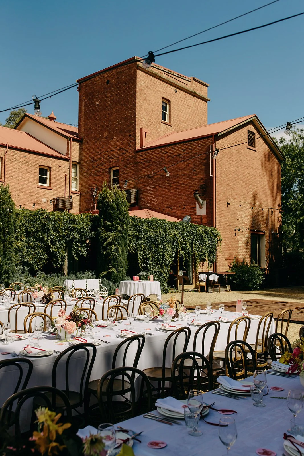 Outdoor dining setup in a garden with a large brick building in the background, tables with white tablecloths, floral centerpieces, chairs, and string lights.
