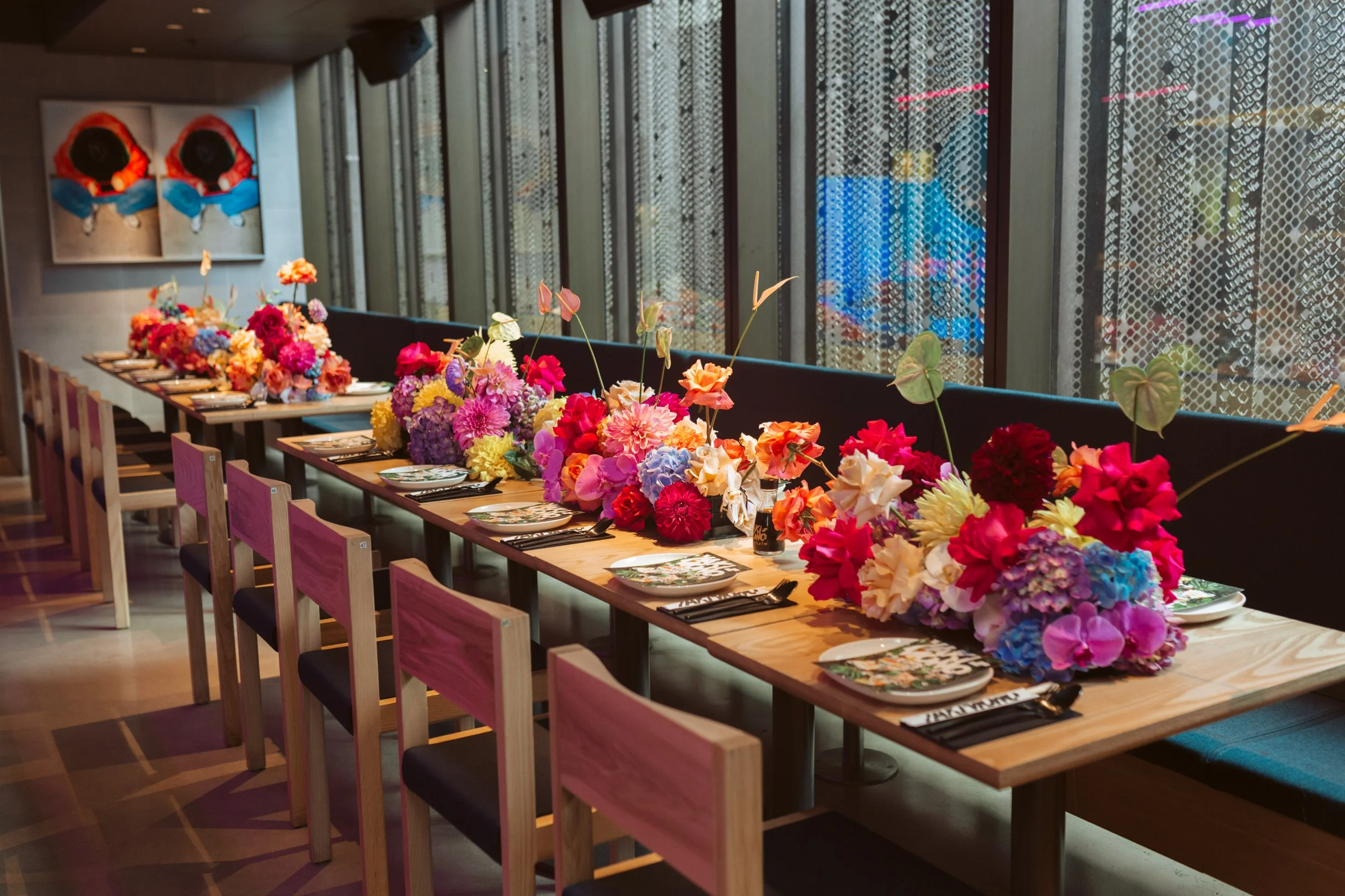 A long dining table decorated with colorful flower arrangements, set in a modern indoor space with large windows and artwork on the wall.