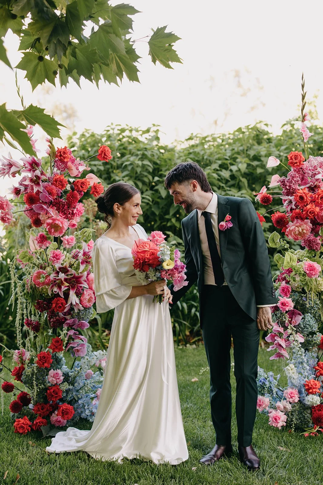 A bride and groom exchanging vows outdoors, surrounded by vibrant pink, red, and purple flowers, with greenery in the background.
