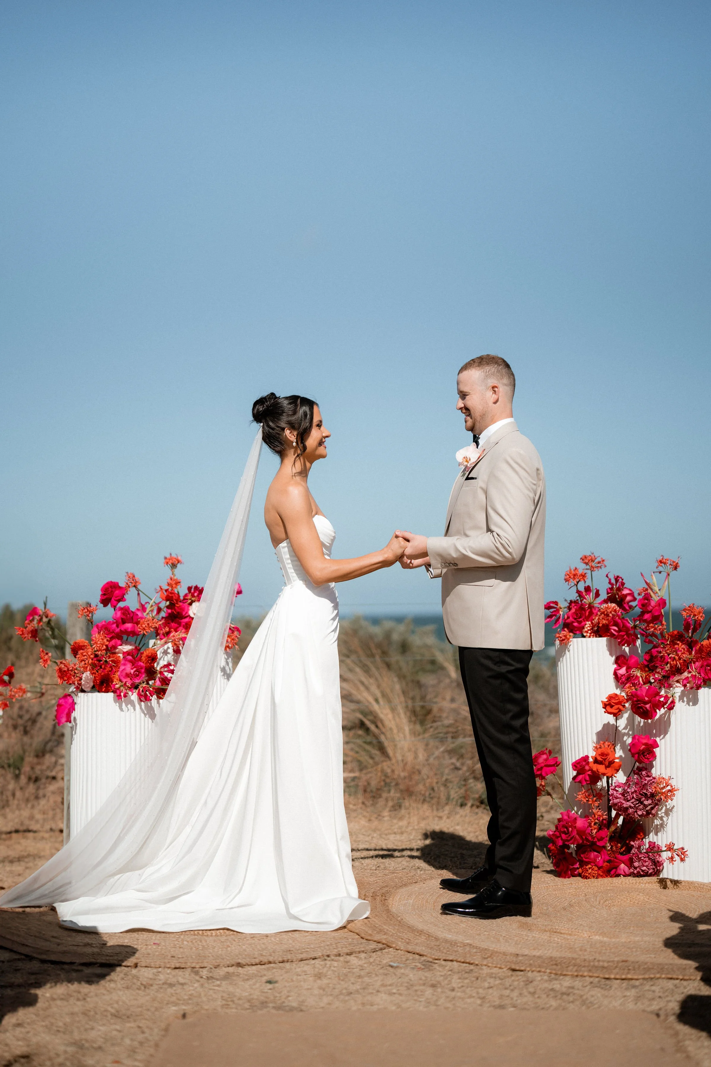 A bride and groom holding hands during an outdoor wedding ceremony with pink and orange flowers on white planters, clear blue sky, and dry landscape in the background.