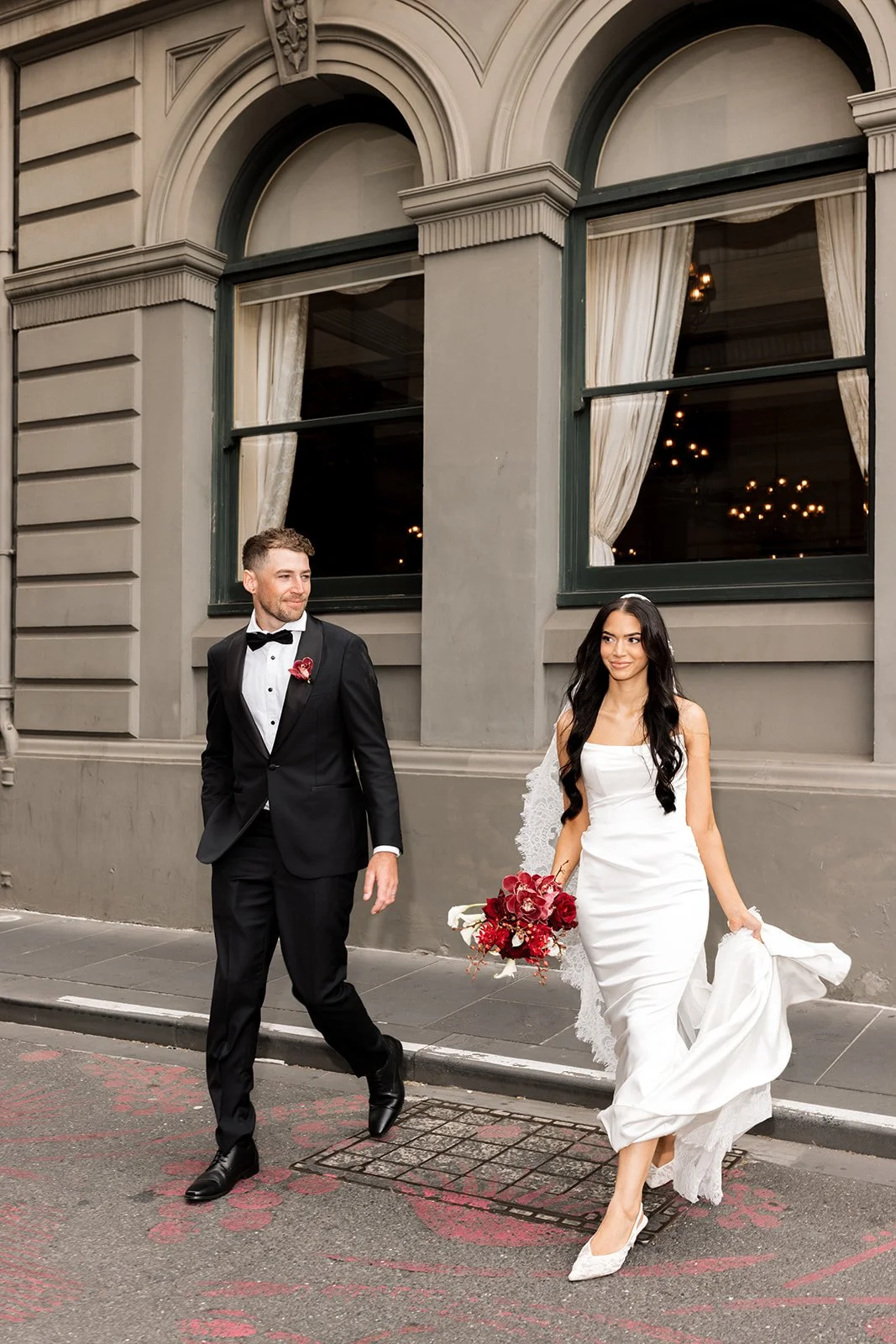 A bride and groom walking on a city street in front of a building with large arched windows, with the bride holding a bouquet of red and white flowers and the groom dressed in a black tuxedo.