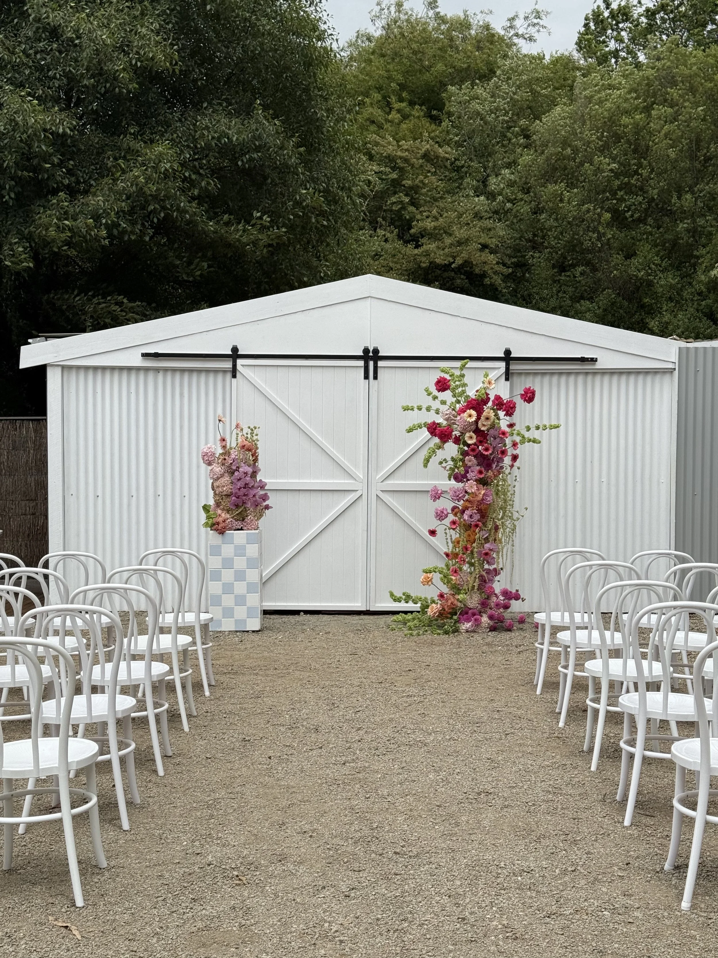 Outdoor wedding ceremony setup with white chairs arranged in rows on a gravel ground, facing a white shed decorated with pink and white flowers arranged around the door, with trees in the background.