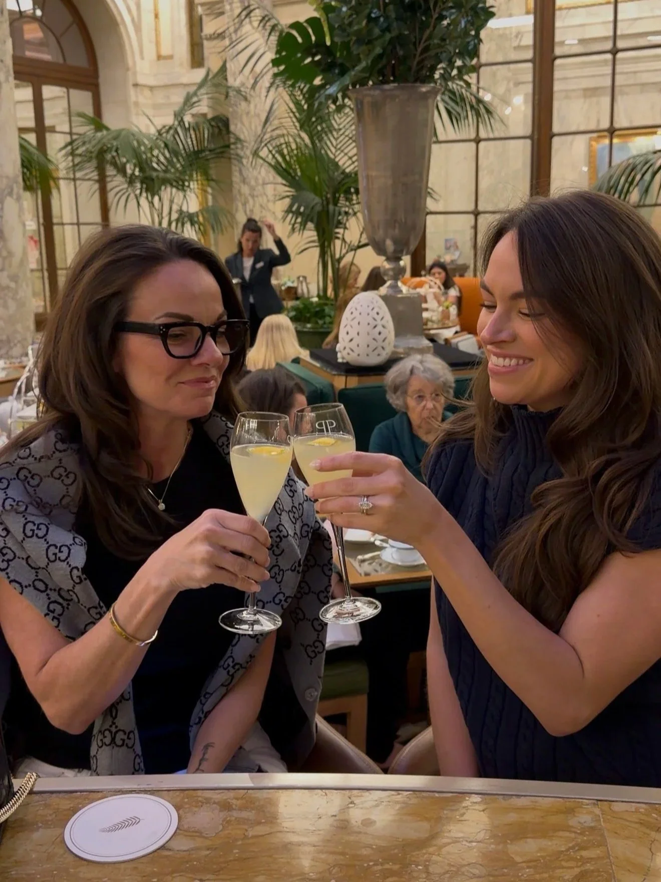 Two women are celebrating with glasses of champagne at a restaurant. The woman on the left has long brown hair, glasses, and is wearing a Gucci scarf. The woman on the right has long wavy brown hair and is smiling. In the background, there are people dining, large plants, and decorative items.