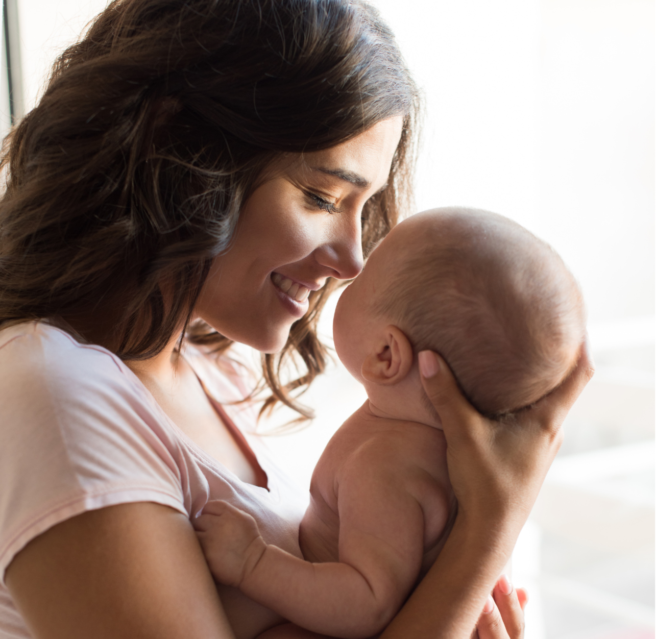 A woman smiling warmly while holding a naked baby close to her face, both inside near a window with soft natural light.