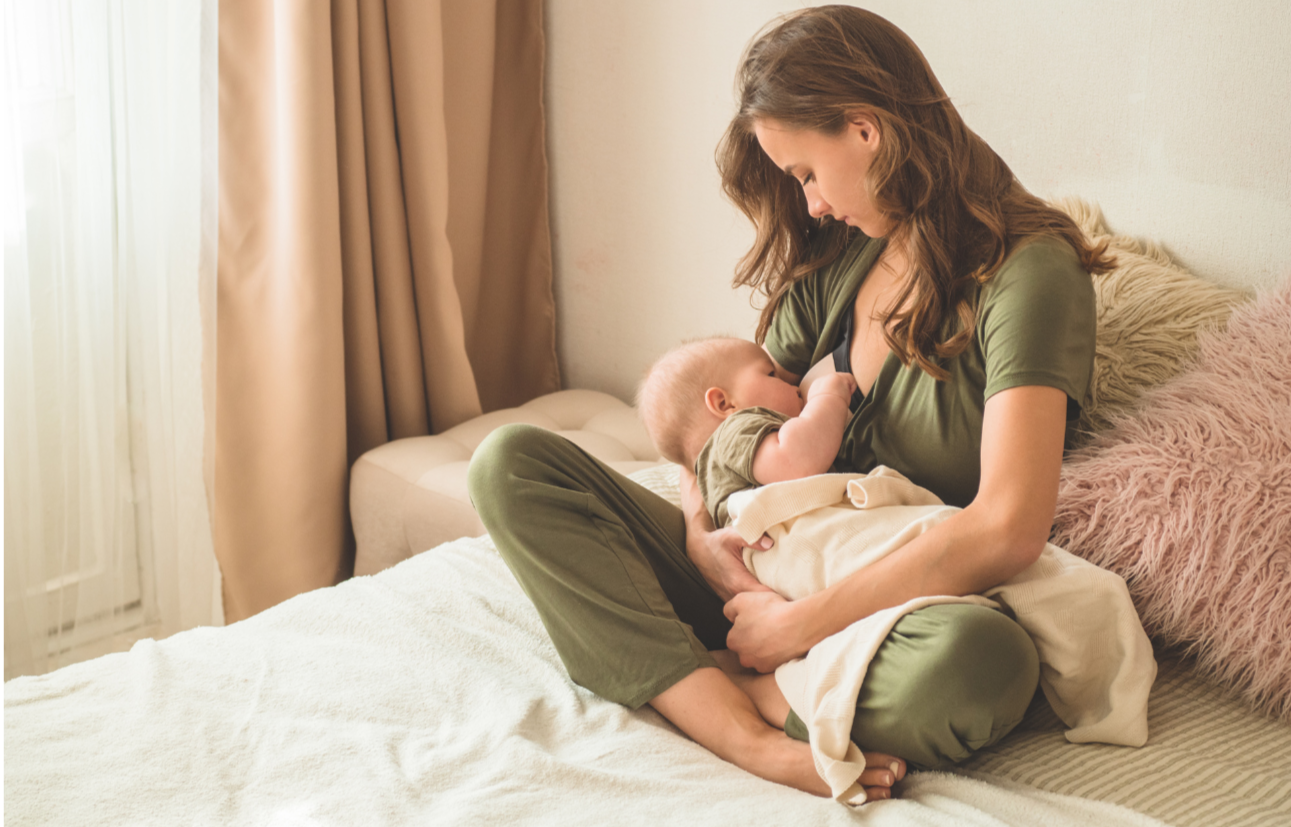 Mother breastfeeding her baby on a bed in a sunlit room with beige curtains and pink fluffy pillows.