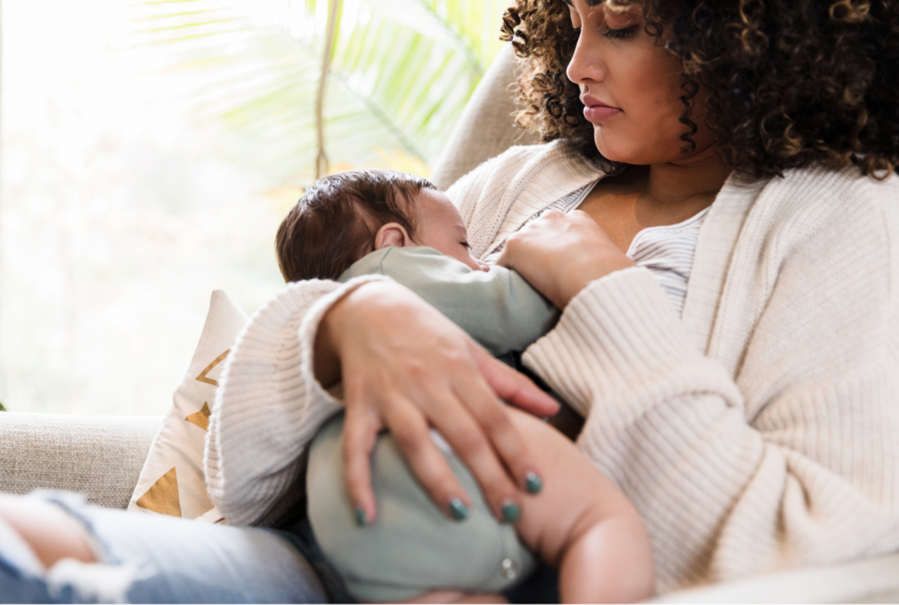 Woman holding a newborn baby on her chest while sitting on a couch near a window with green plants in the background.