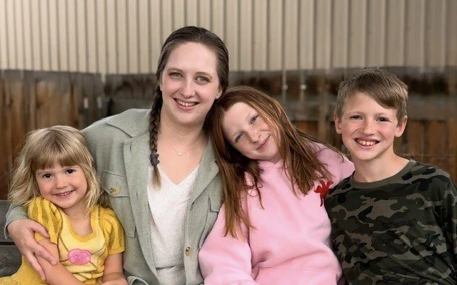 A woman with two girls and a boy smiling together outdoors in a backyard.