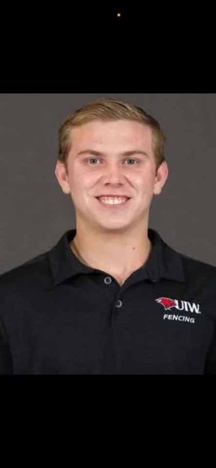 A young man with short blonde hair smiling in front of a gray background, wearing a black polo shirt with the UIW Fencing logo.