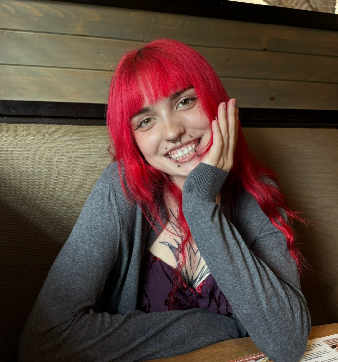 A woman with bright red hair and multiple facial piercings sitting at a table in a restaurant or cafe, smiling and resting her head on her hand.