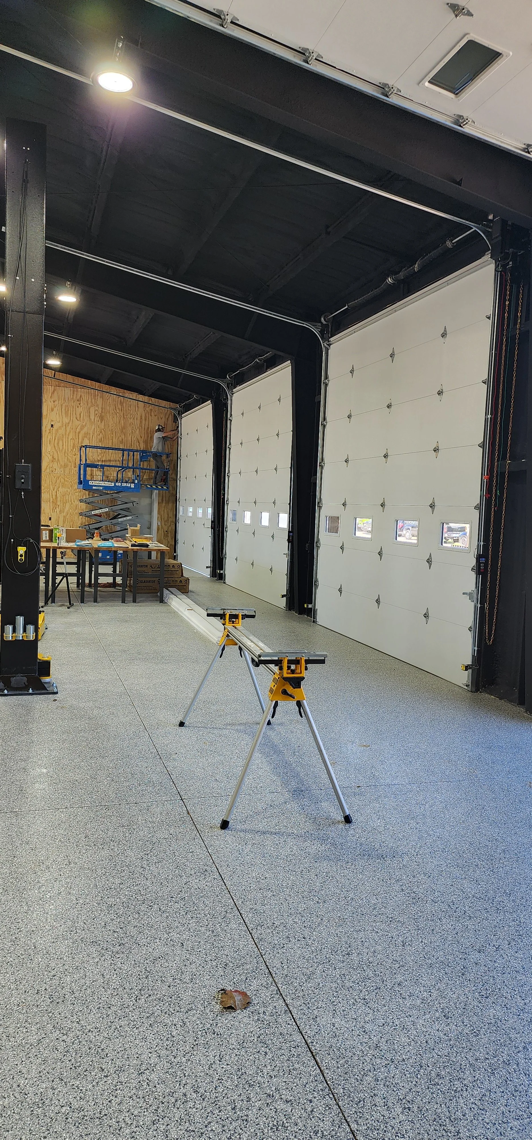 The image shows the interior of a garage or workshop with two large white garage doors, one partially open. There is a worktable with tools and materials, a person on a scissor lift working on the wall, and a portable workbench with a saw on it. The space has a gray speckled floor, black ceiling and wall accents, and wooden paneling on the far wall.