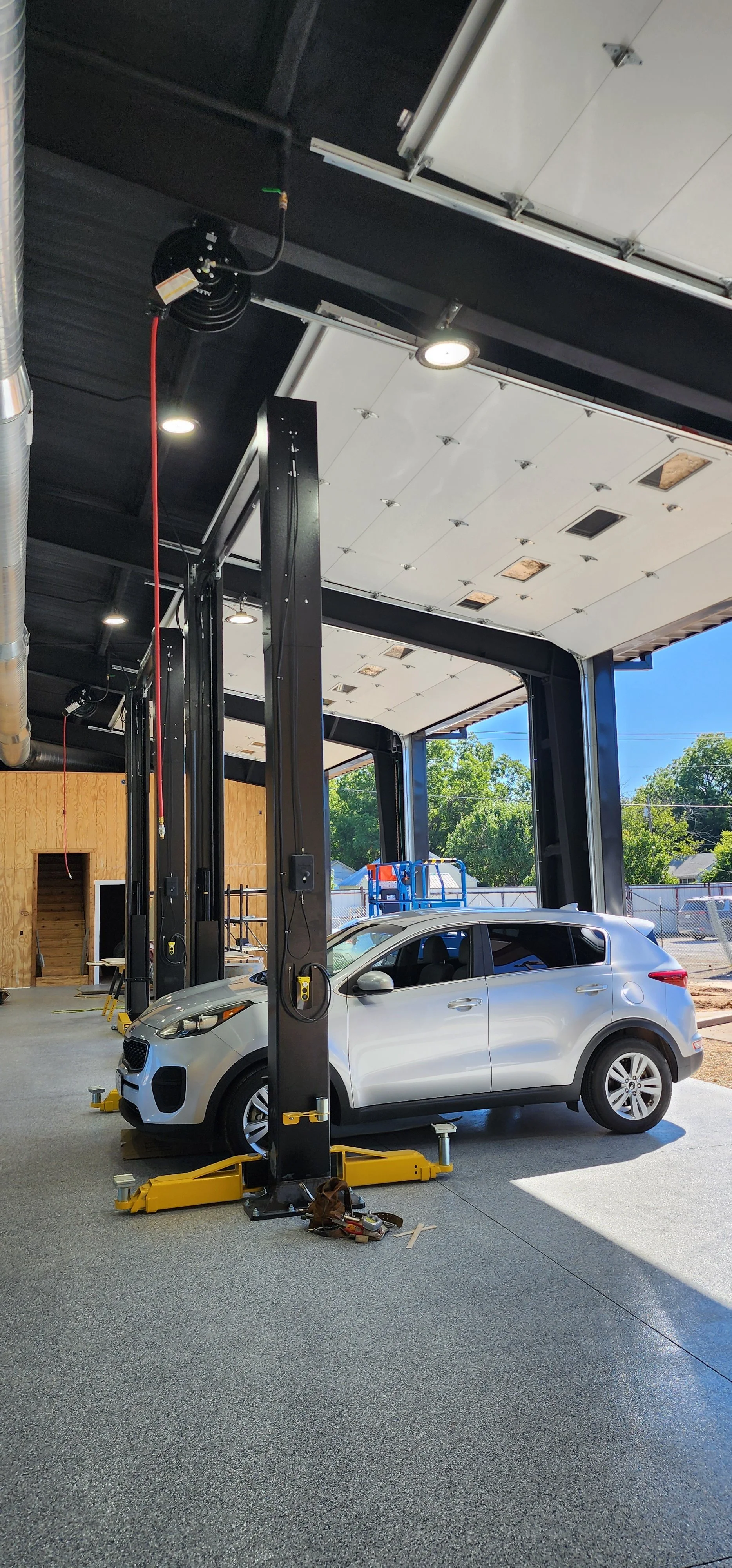 Interior of an auto repair shop or garage with a silver car lifted on hydraulic lifts, with tools and equipment on the floor, and a wooden wall in the background.