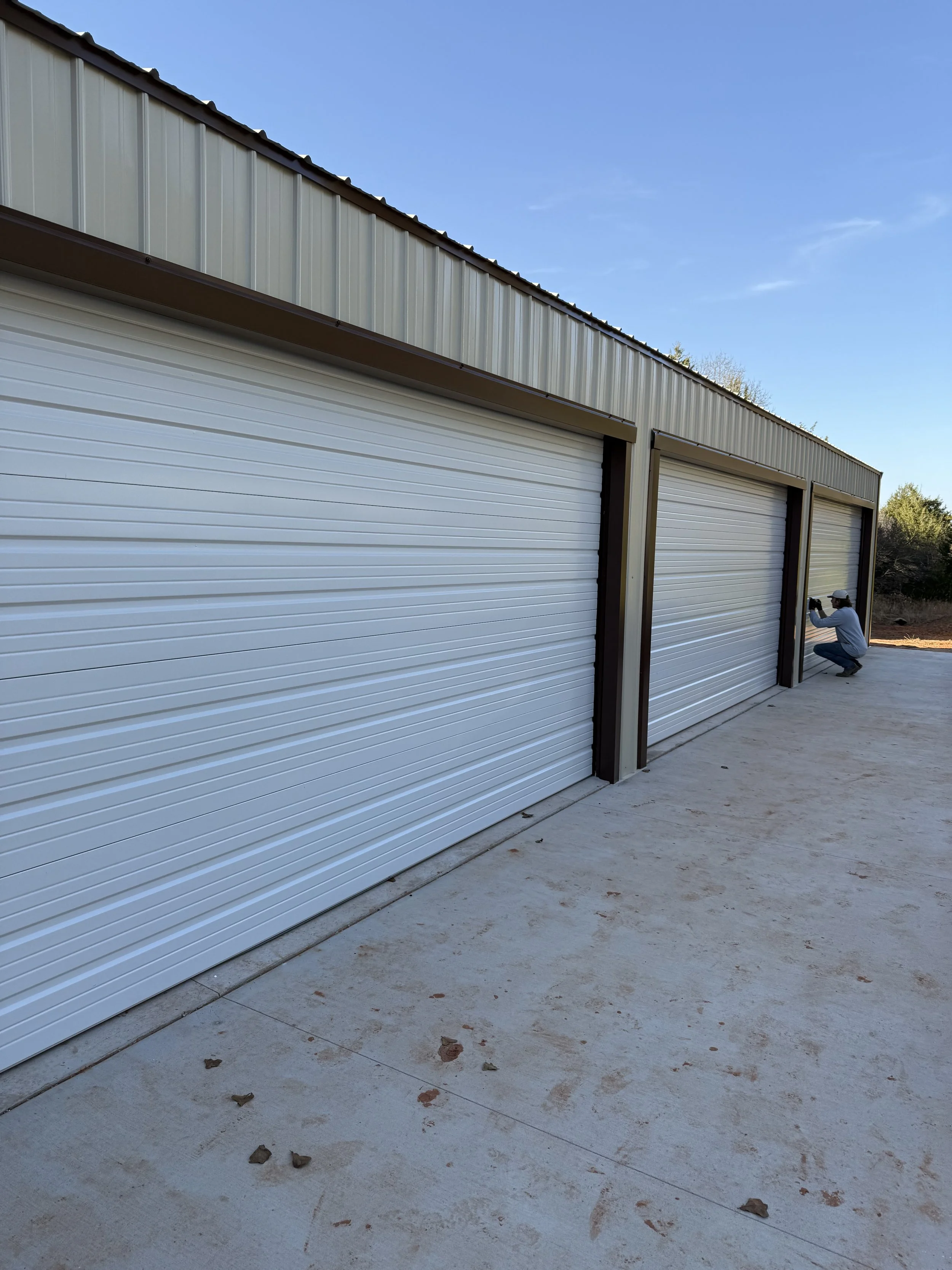 Exterior view of a large metal storage units with white roll-up doors on a concrete ground, with a person squatting and taking a photo near one of the doors, under a clear blue sky.