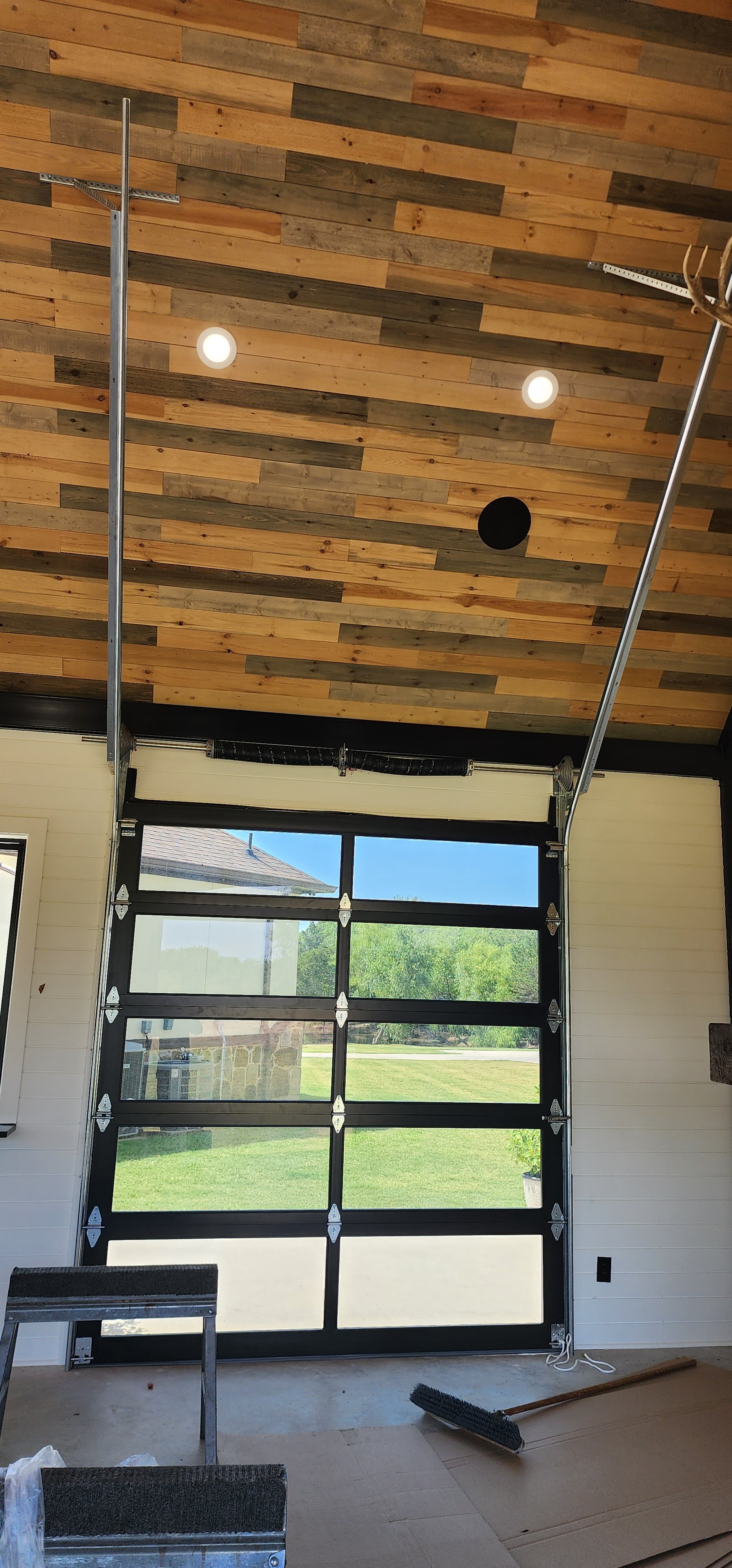 Interior view of a garage with a wooden plank ceiling, large glass garage door with black framing, and a view of green trees outside. The space appears to be under construction or renovation.