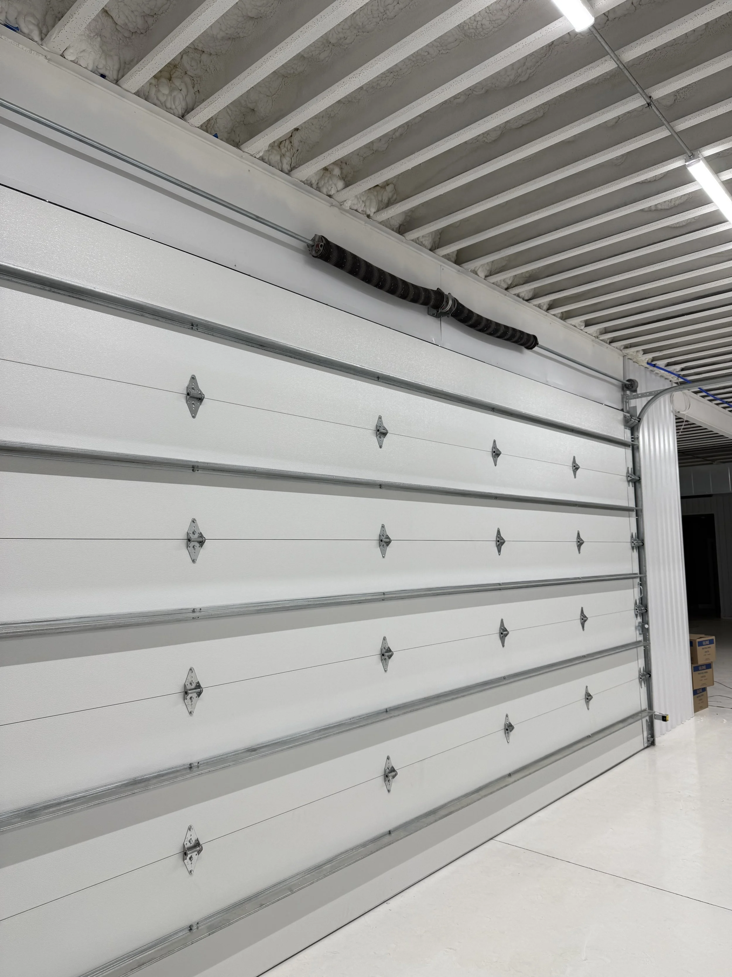 An industrial or warehouse setting with a white rolling metal door and a black cable or conduit running along the top of the door. The ceiling above has insulation between beams, and there's a fluorescent light fixture visible.