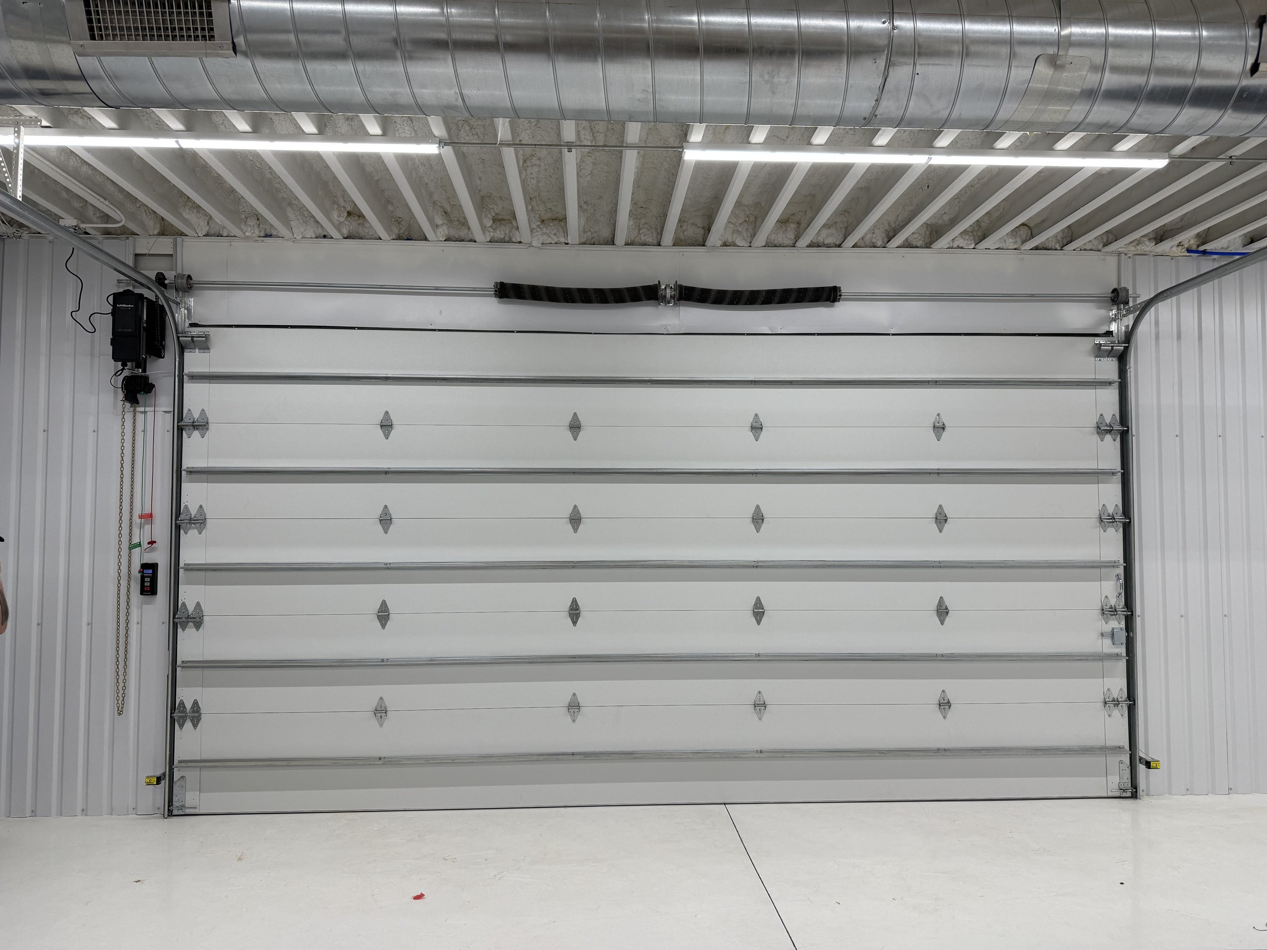 White garage door inside a spacious industrial garage with metal walls, ceiling insulation, and ceiling-mounted LED lights.