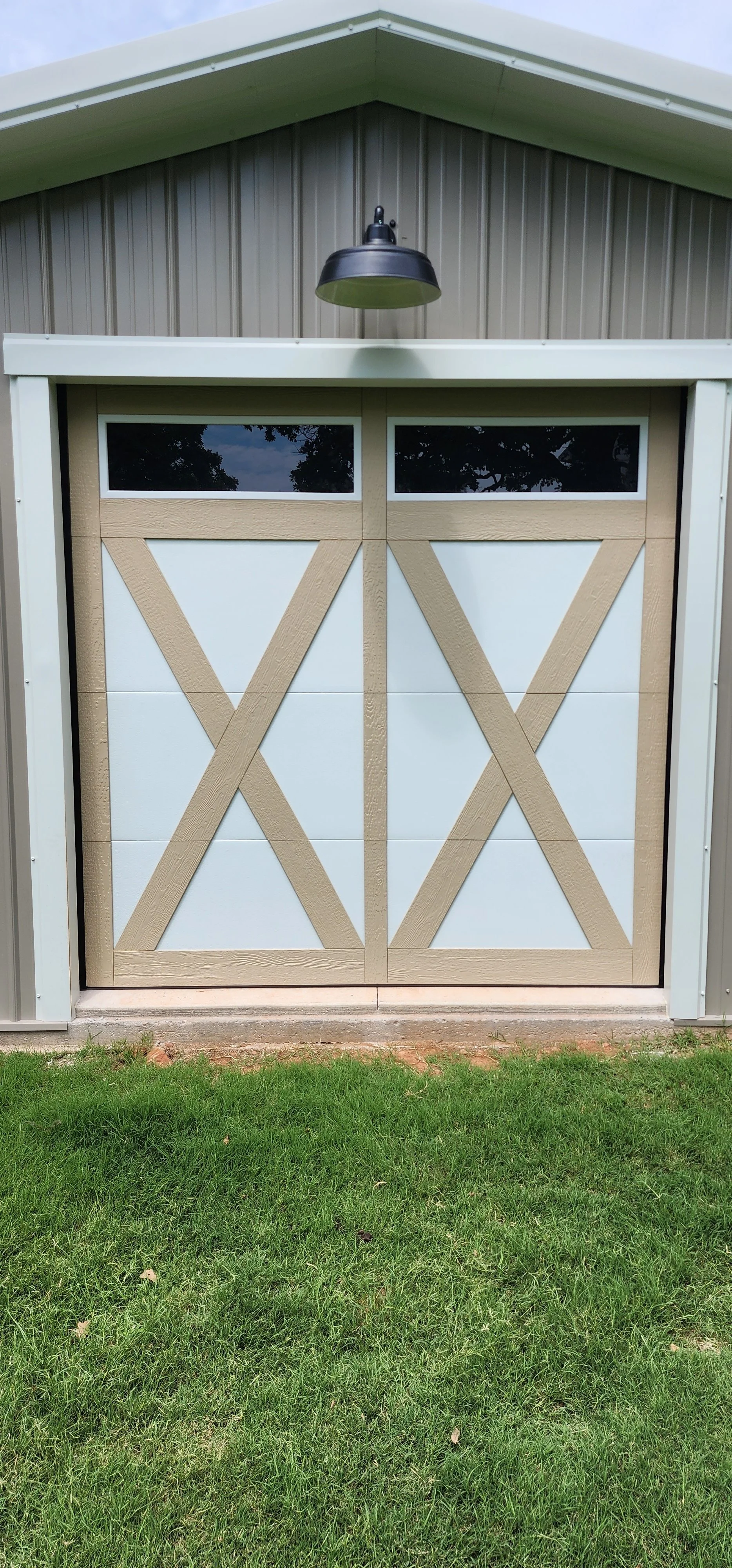 A garage door designed to resemble a barn door with beige faux wood cross beams and white panels, set against a metal building with a light gray roof. There is a black outdoor light fixture above the door, and a grassy yard in front.