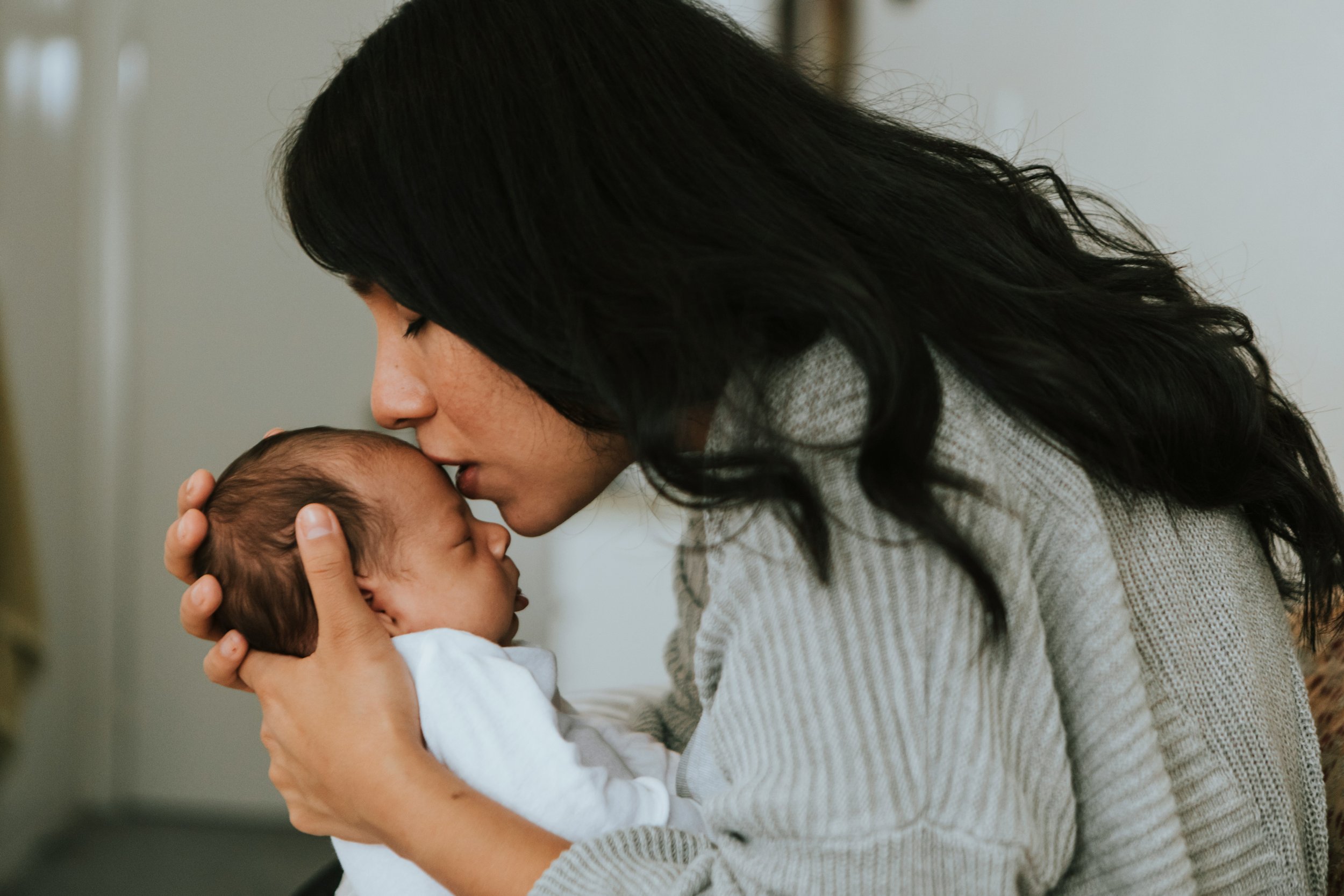 A woman with long dark hair gently pressing her forehead to a baby's forehead, both with closed eyes, in a tender moment indoors.