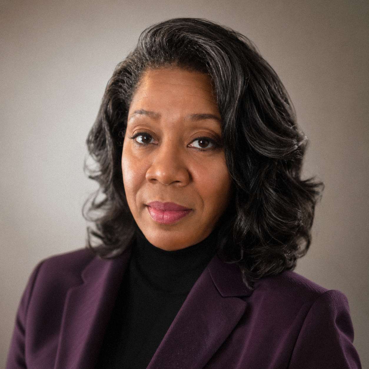 Close-up portrait of an African American woman with shoulder-length curly black hair, wearing a dark purple blazer and black turtleneck, against a neutral background.