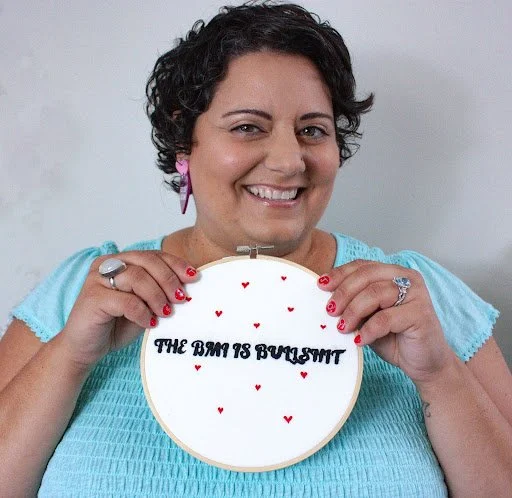 Woman smiling and holding an embroidered hoop with the words "fuck diet culture" and small red hearts.