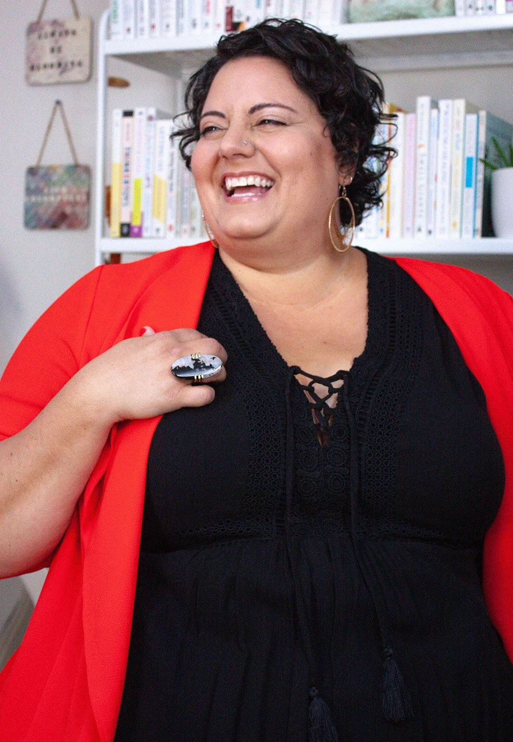 Sophia Apostol,  the founder of Fat Joy Creation Lab, and a joyful, plus-size white woman with curly brown hair, beams in a vibrant red jacket over a black dress, radiating body positivity and empowerment against a backdrop of colorful bookshelves.