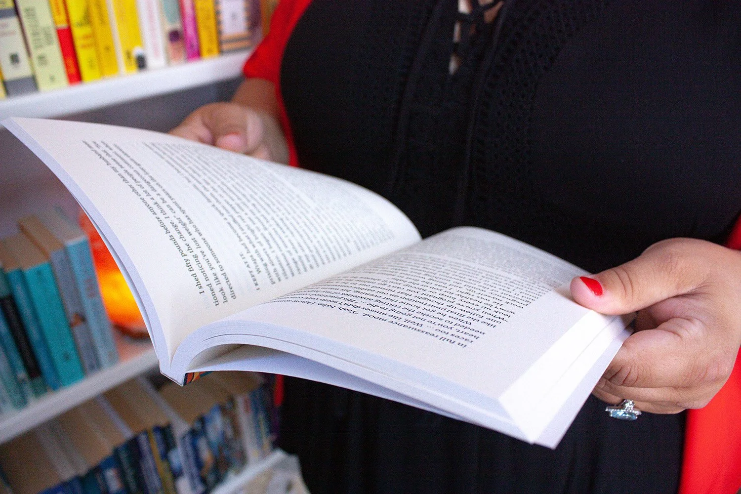 Sophia Apostol,  the founder of Fat Joy Creation Lab and a white woman with a bold red shawl turns the pages of a book, against a vibrant backdrop of colorful shelves. A celebration of body positivity and creativity shines through.